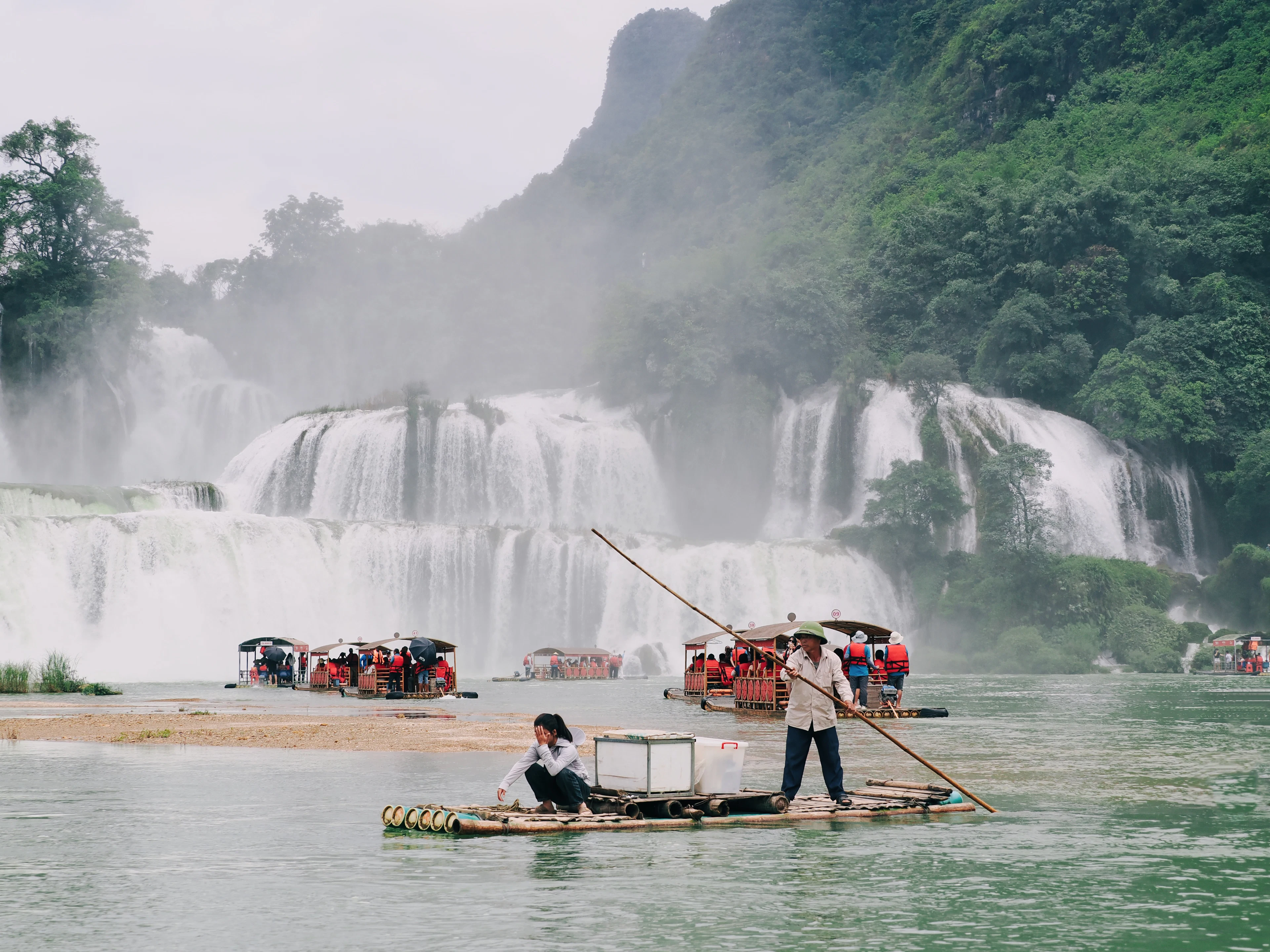 Bamboo rafts on green water beneath a wide, tiered waterfall among limestone mountains