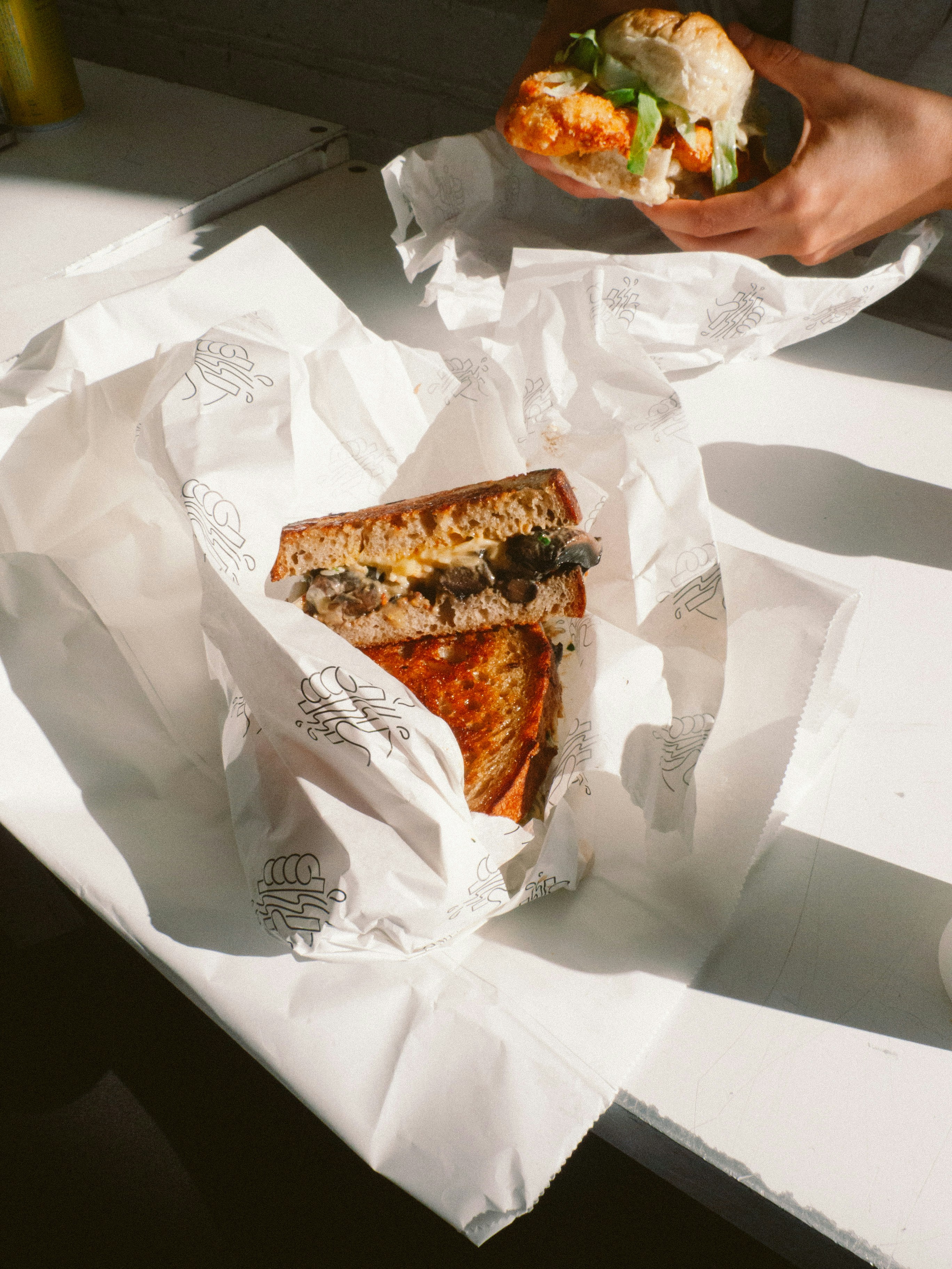 Hands holding a sandwich, another on a sunlit table.