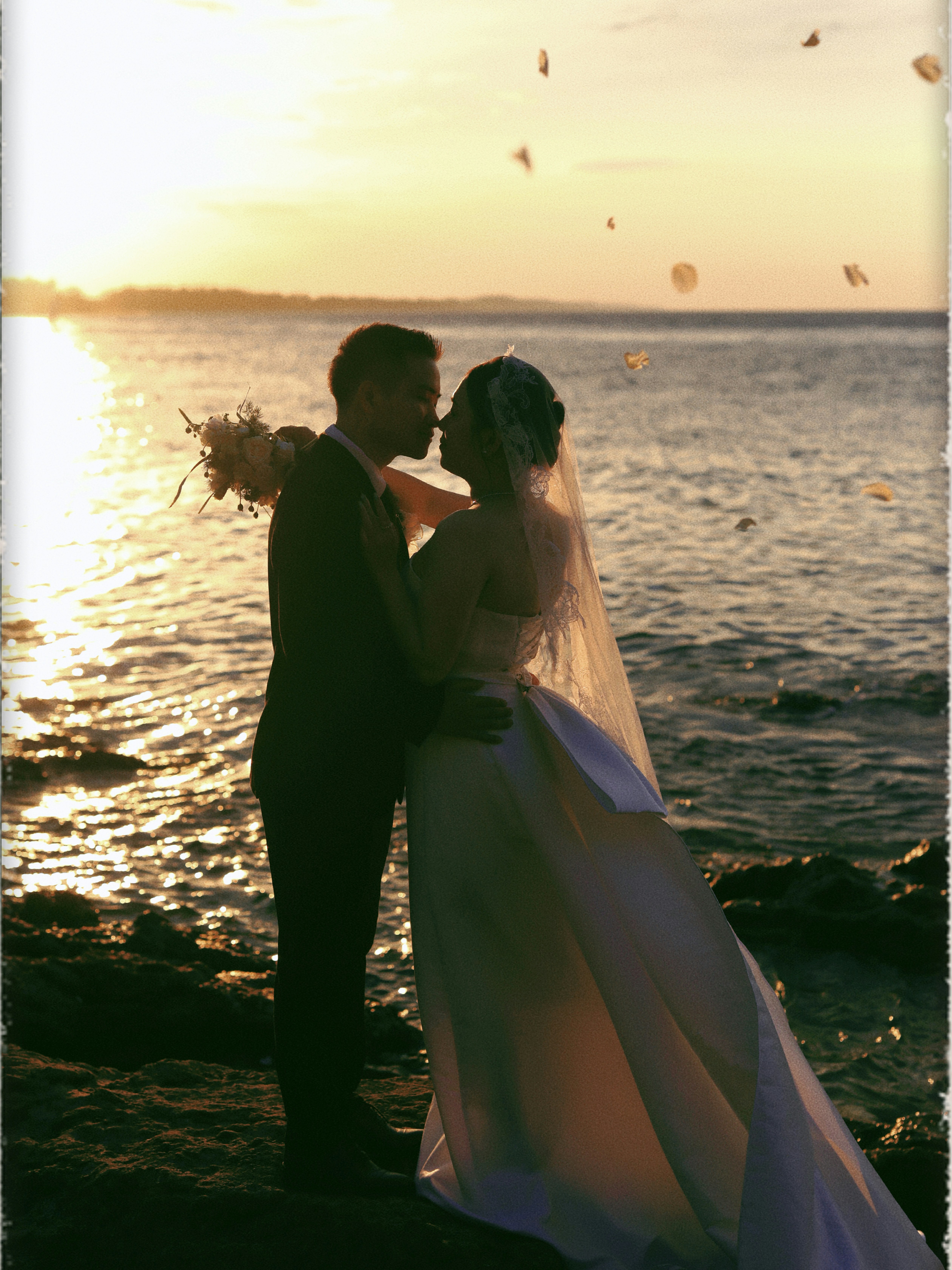 Silhouetted bride and groom embrace by the ocean at sunset.
