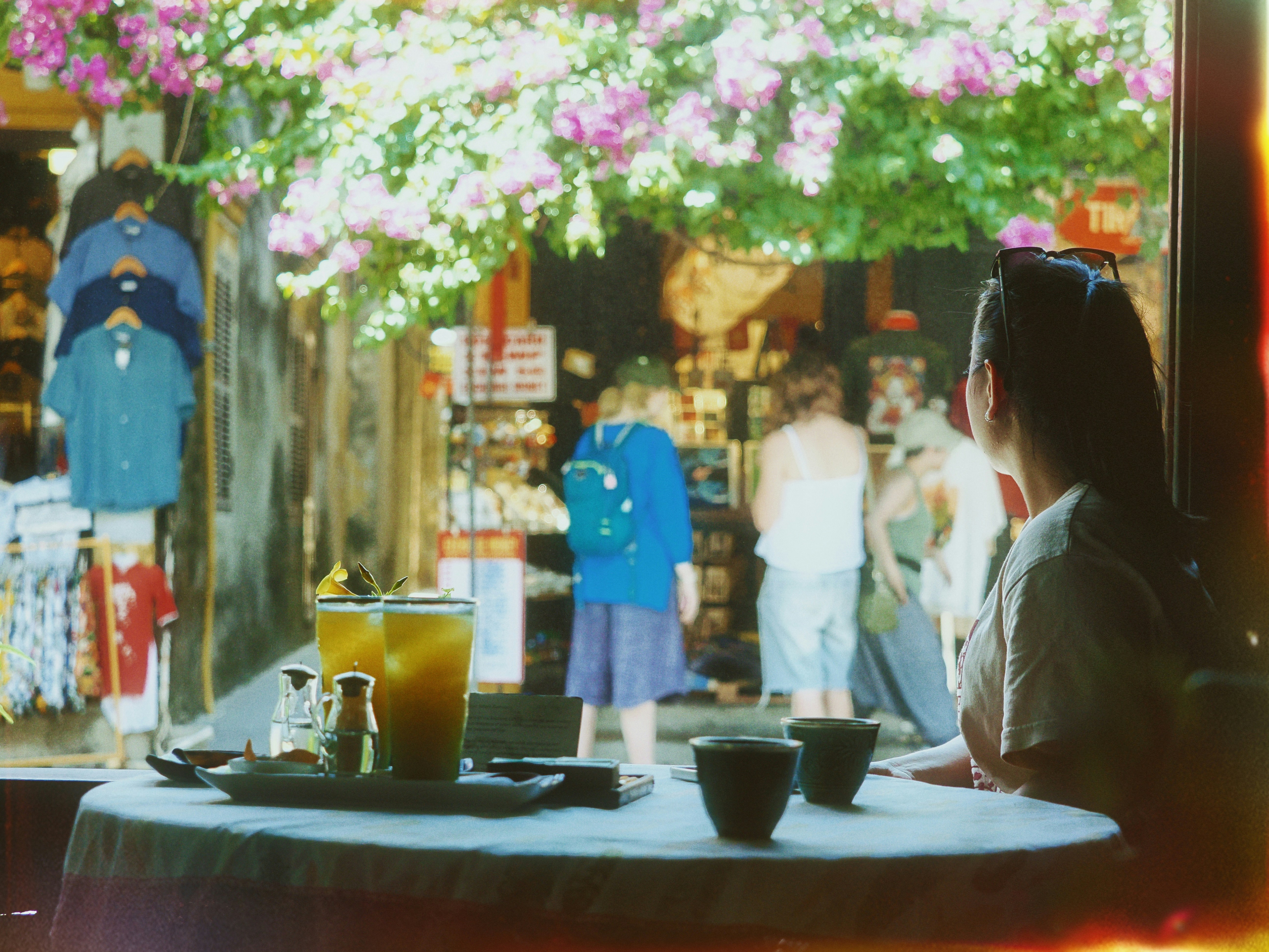 Woman at cafe table watching street with shops and flowers.