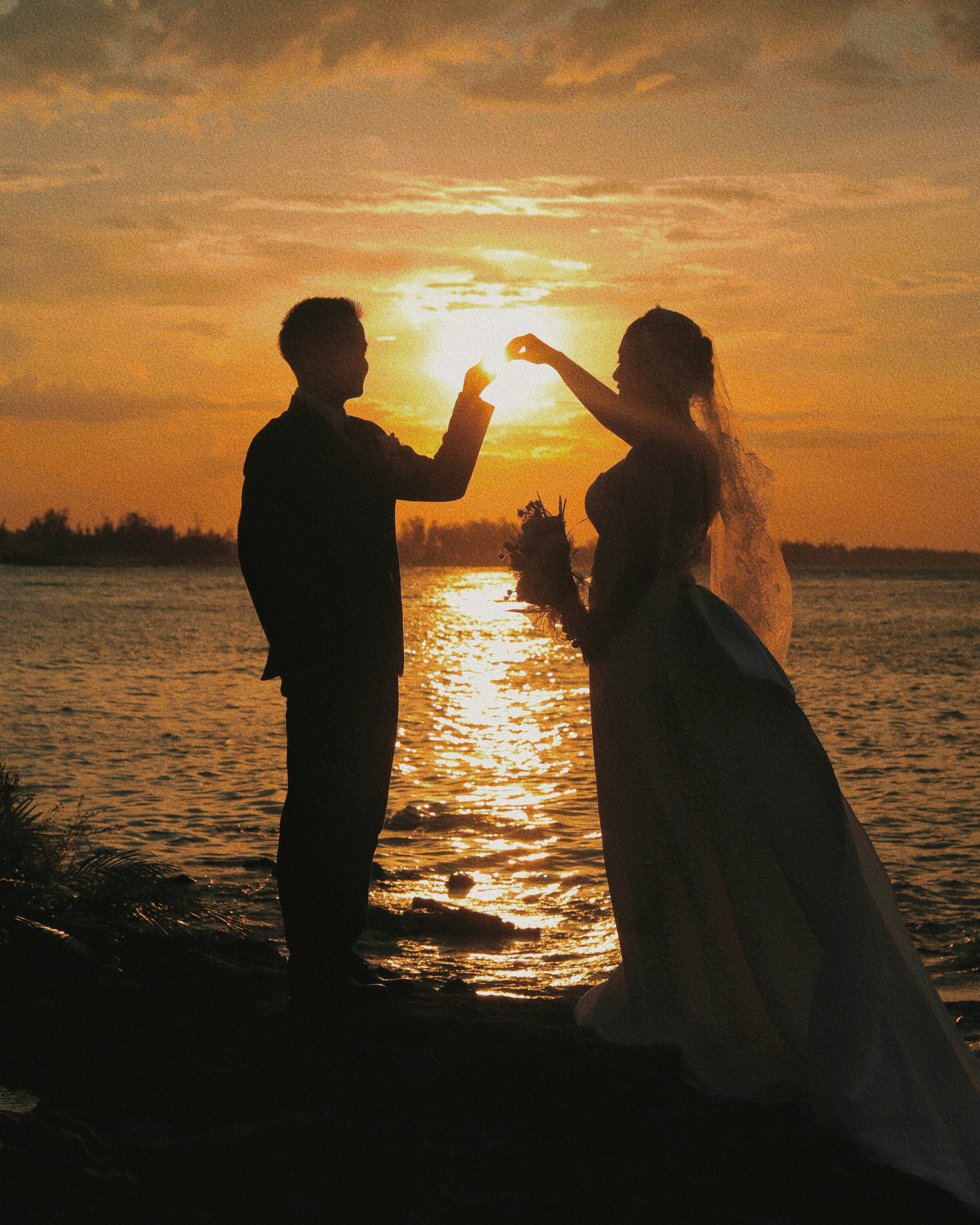 Silhouetted wedding couple holding the setting sun over water.