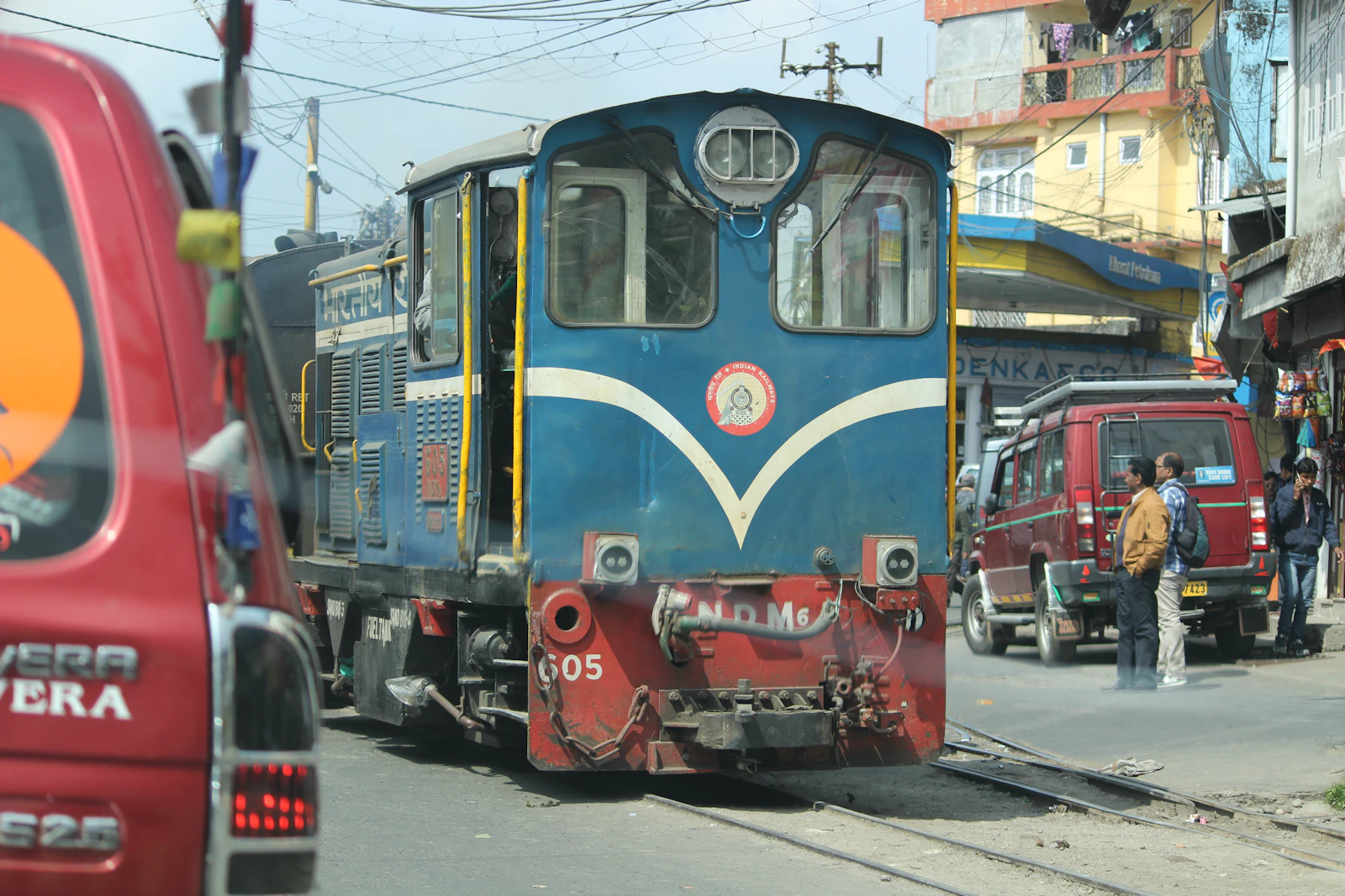A blue Darjeeling Himalayan Railway locomotive edging along a hill bazaar street between jeeps, shopfronts and people