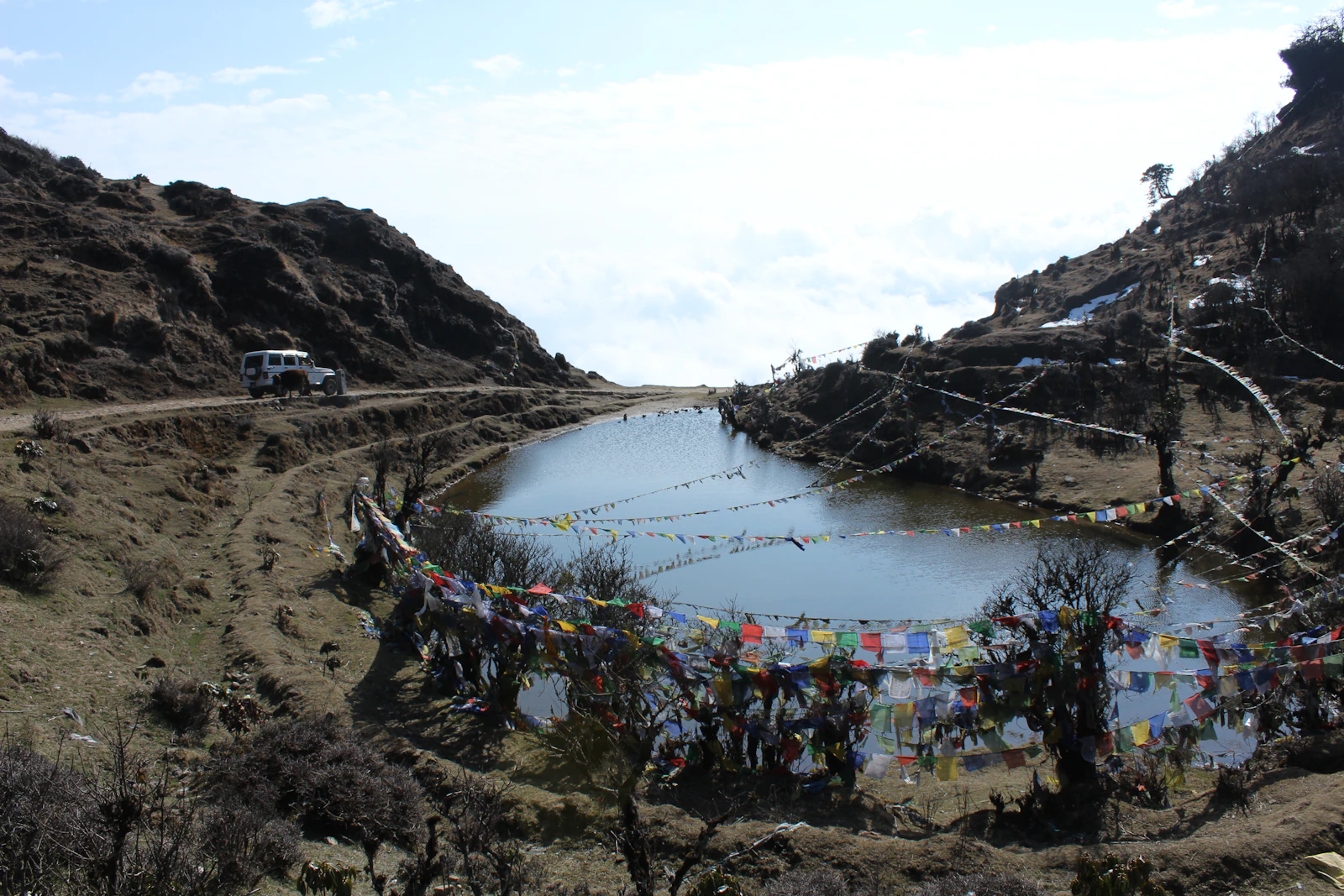 A white four wheel drive on a rough track above a high mountain pond ringed with prayer flags, cloud below