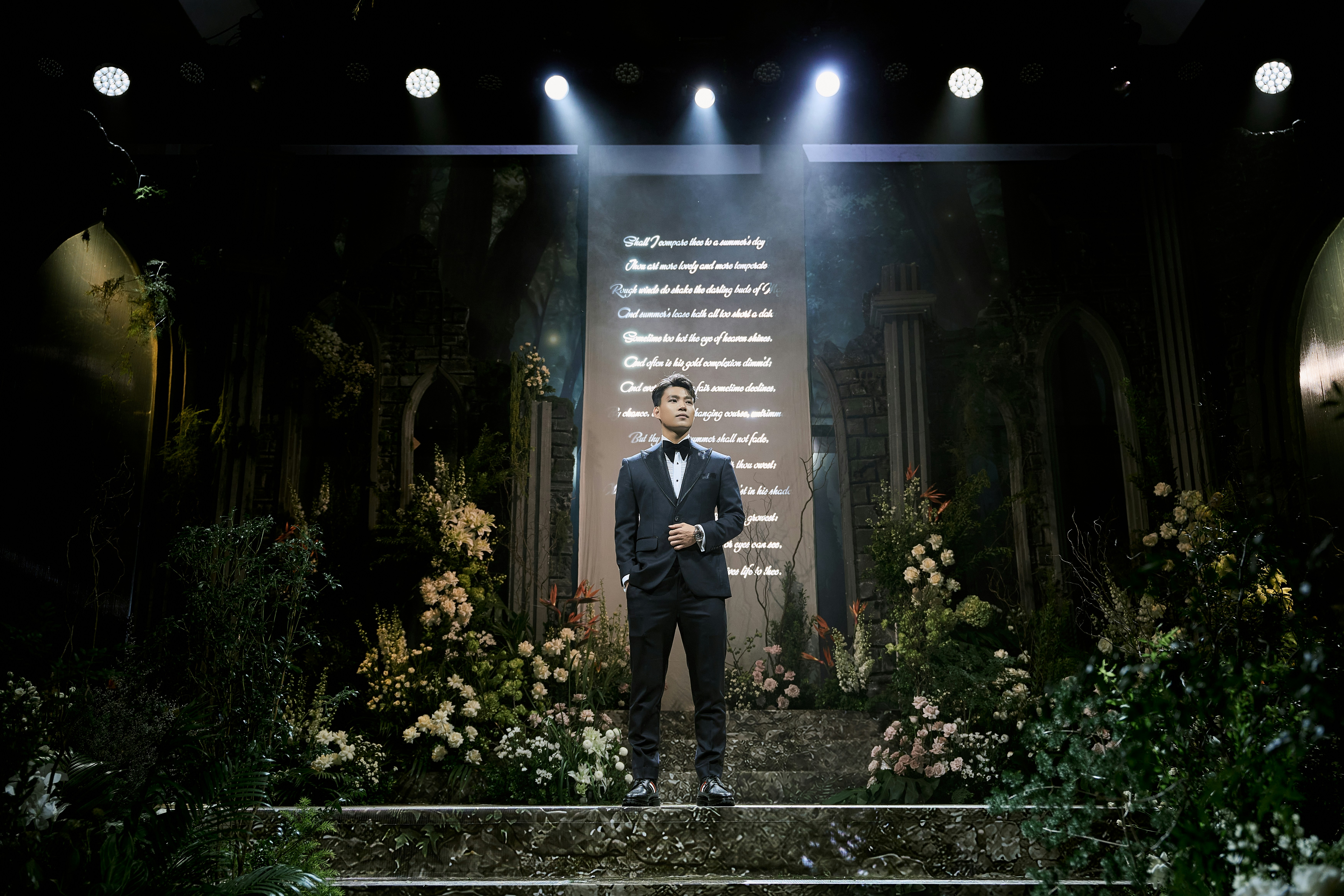 Man in tuxedo on stage with lit screen and flowers