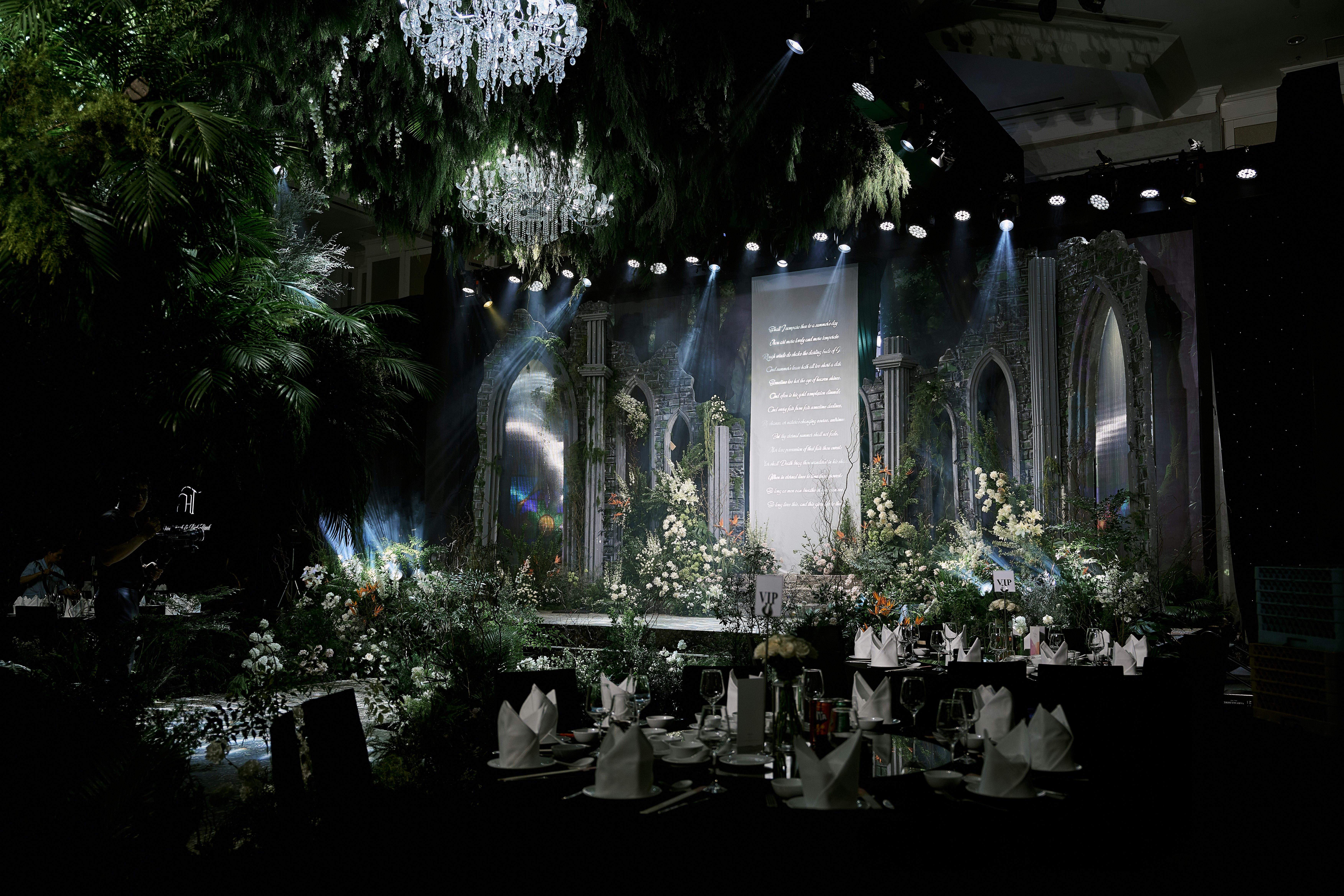 Elegant event stage with white flowers, chandeliers, and dramatic lighting.