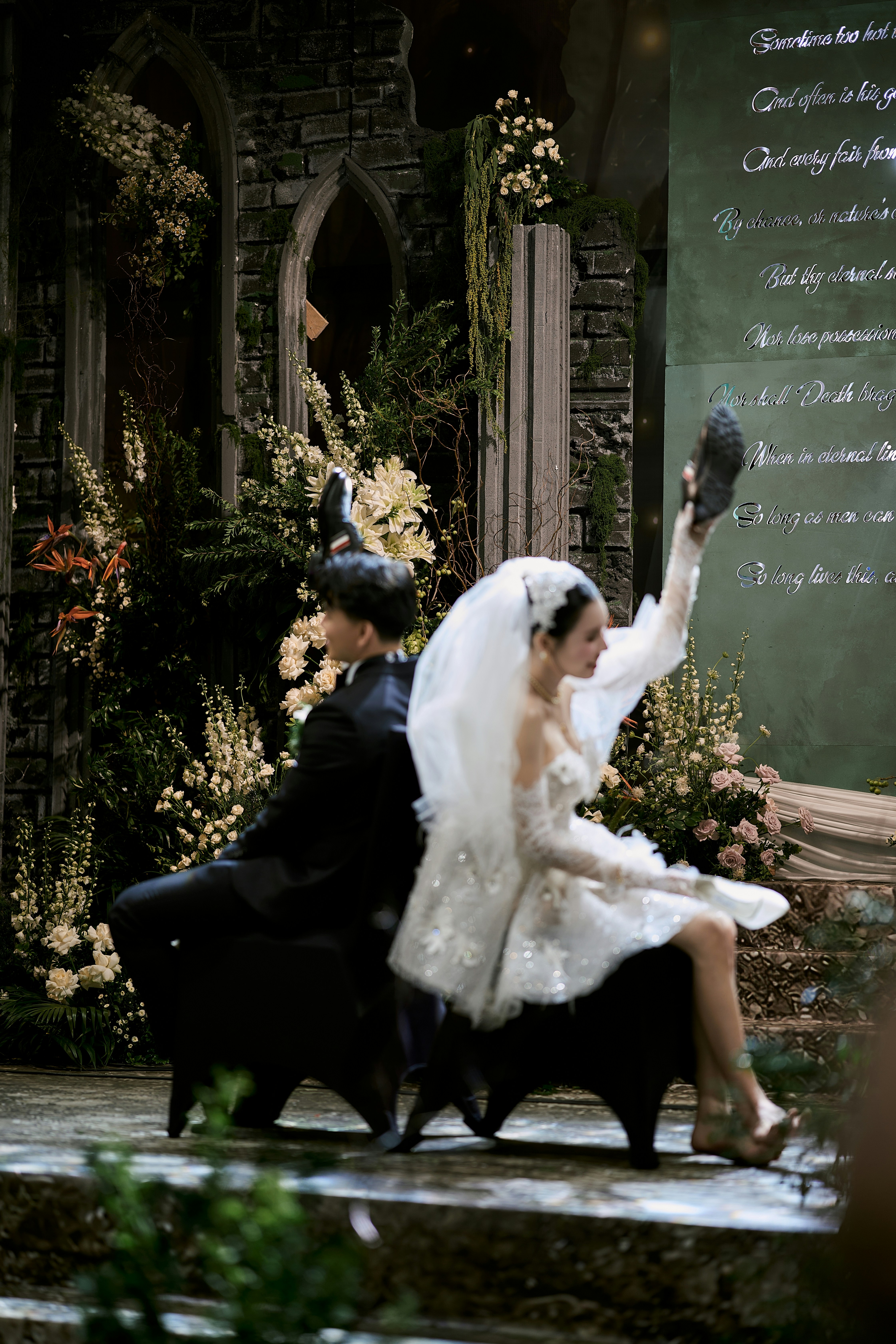 Bride and groom sit back-to-back, bride holds up a shoe