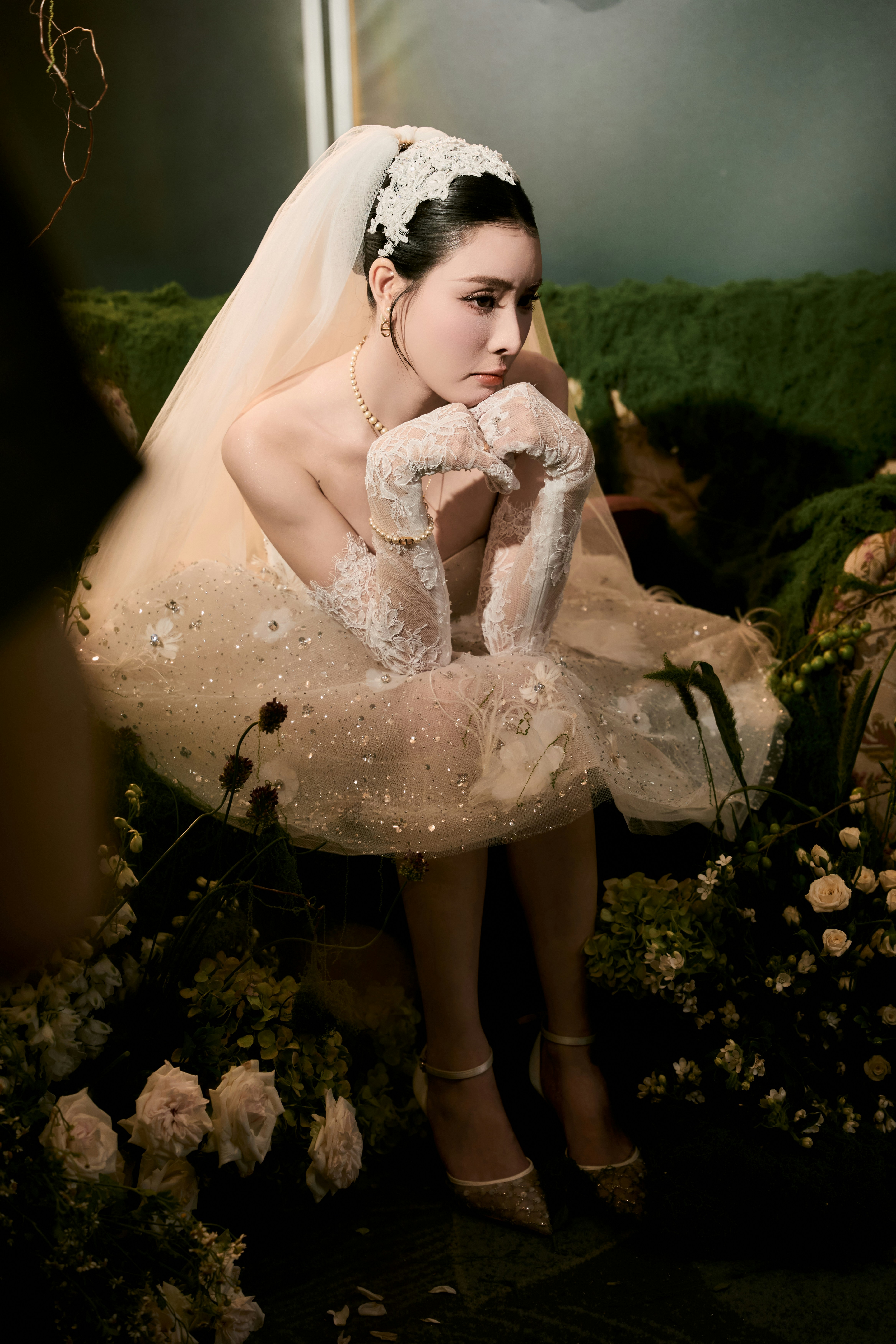 Pensive bride in wedding dress sits among flowers and moss
