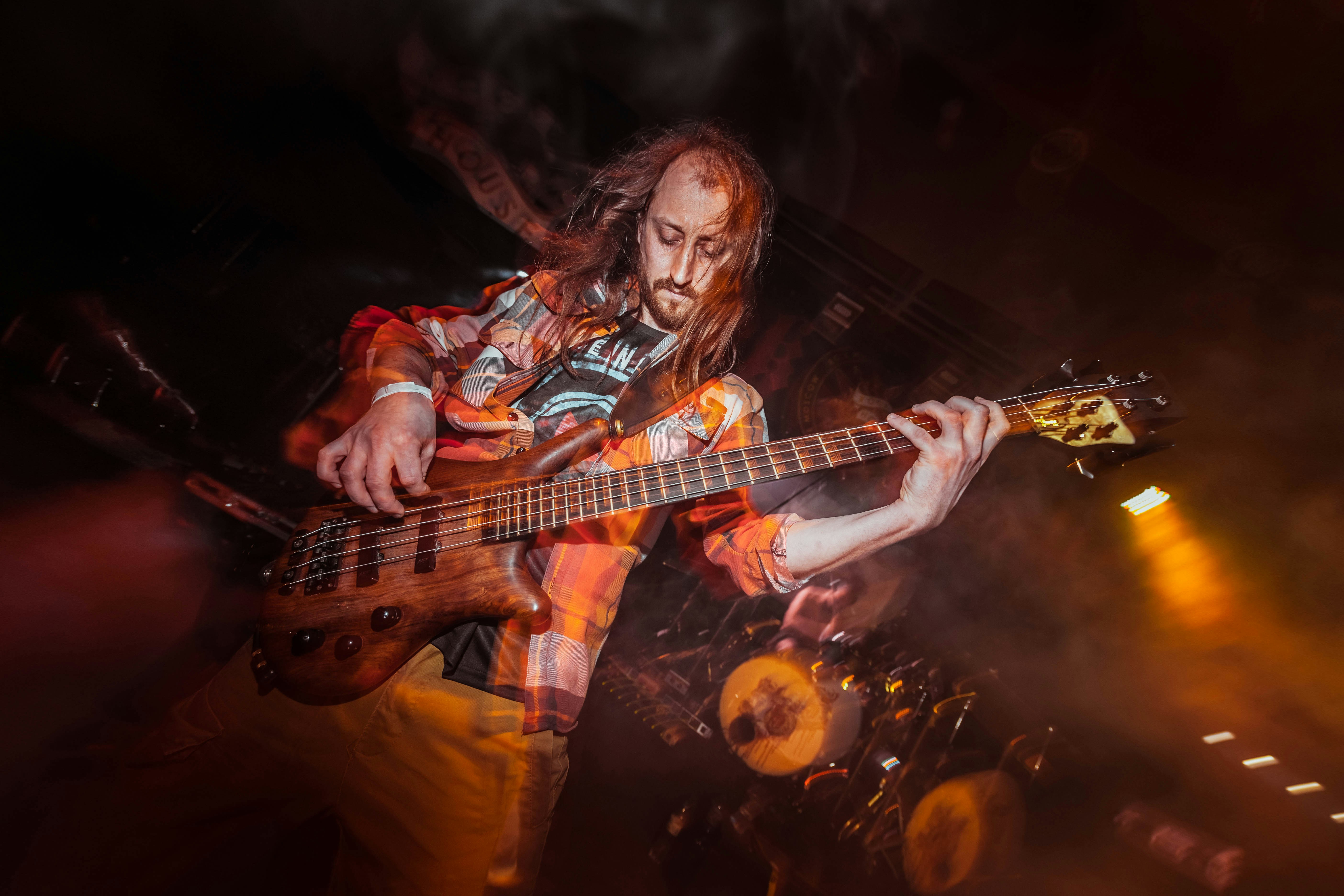 A bearded musician plays bass guitar on a dark stage.