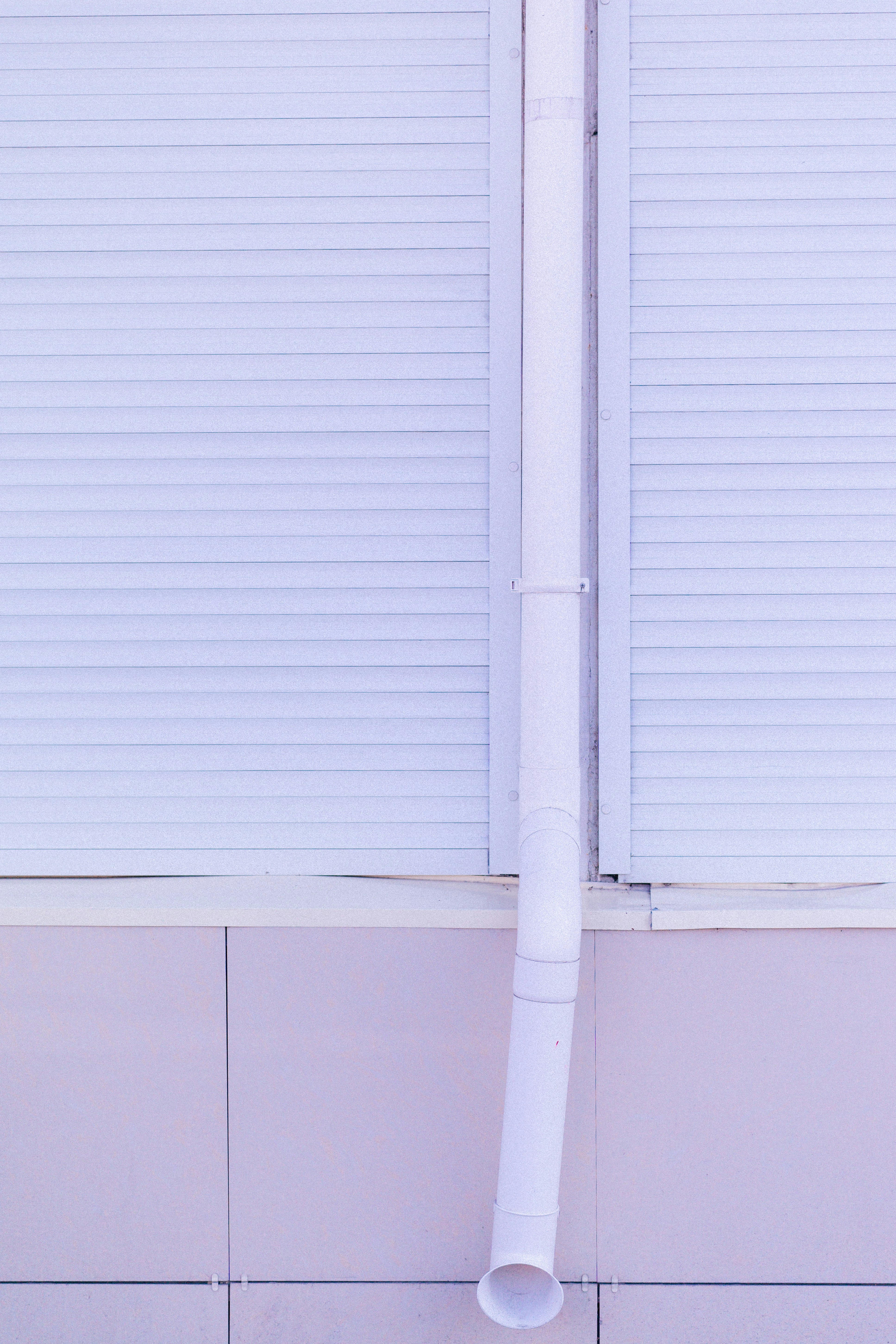 White downspout on a building with light purple roller shutters.