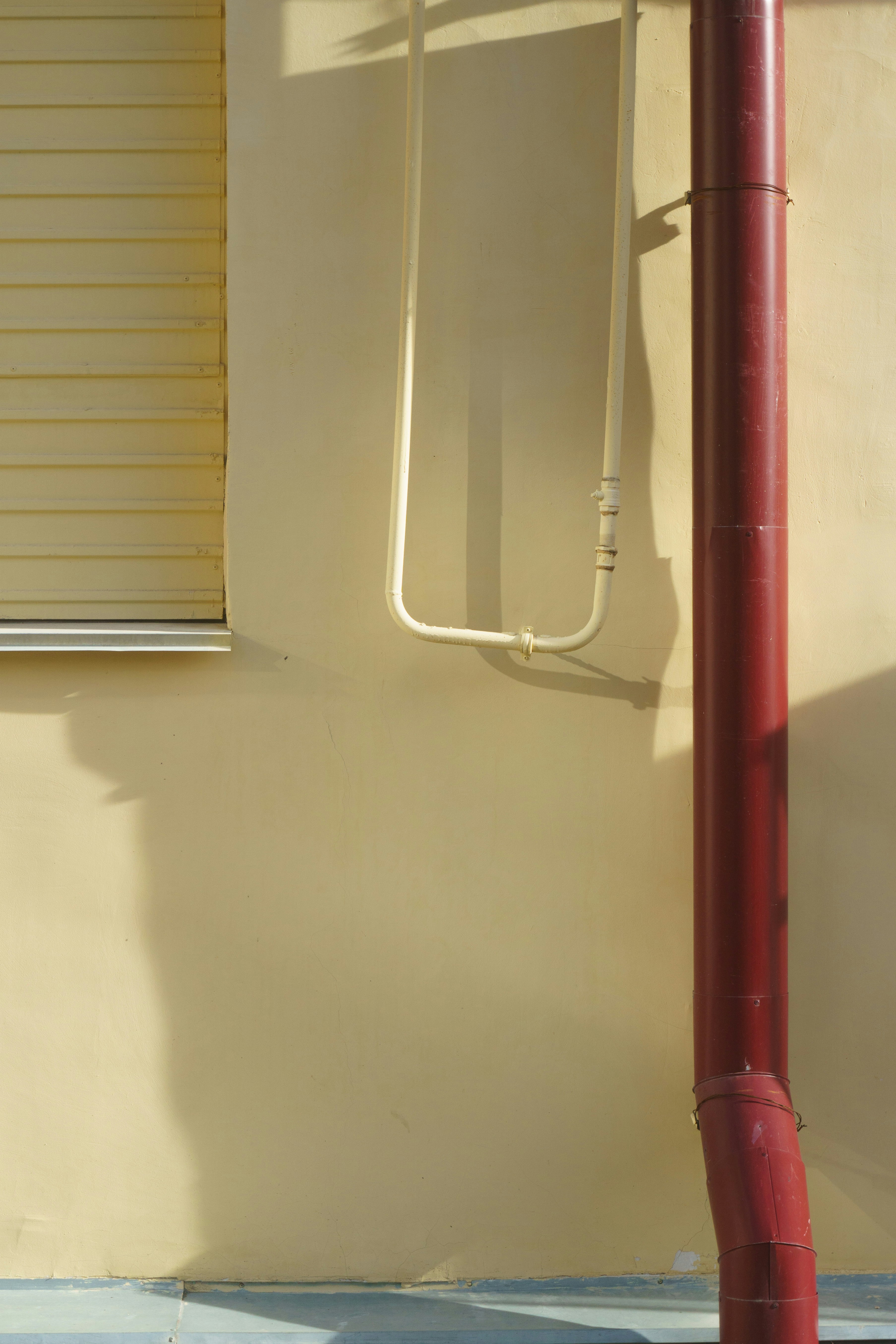 A beige wall with a red downspout, window, and pipes.