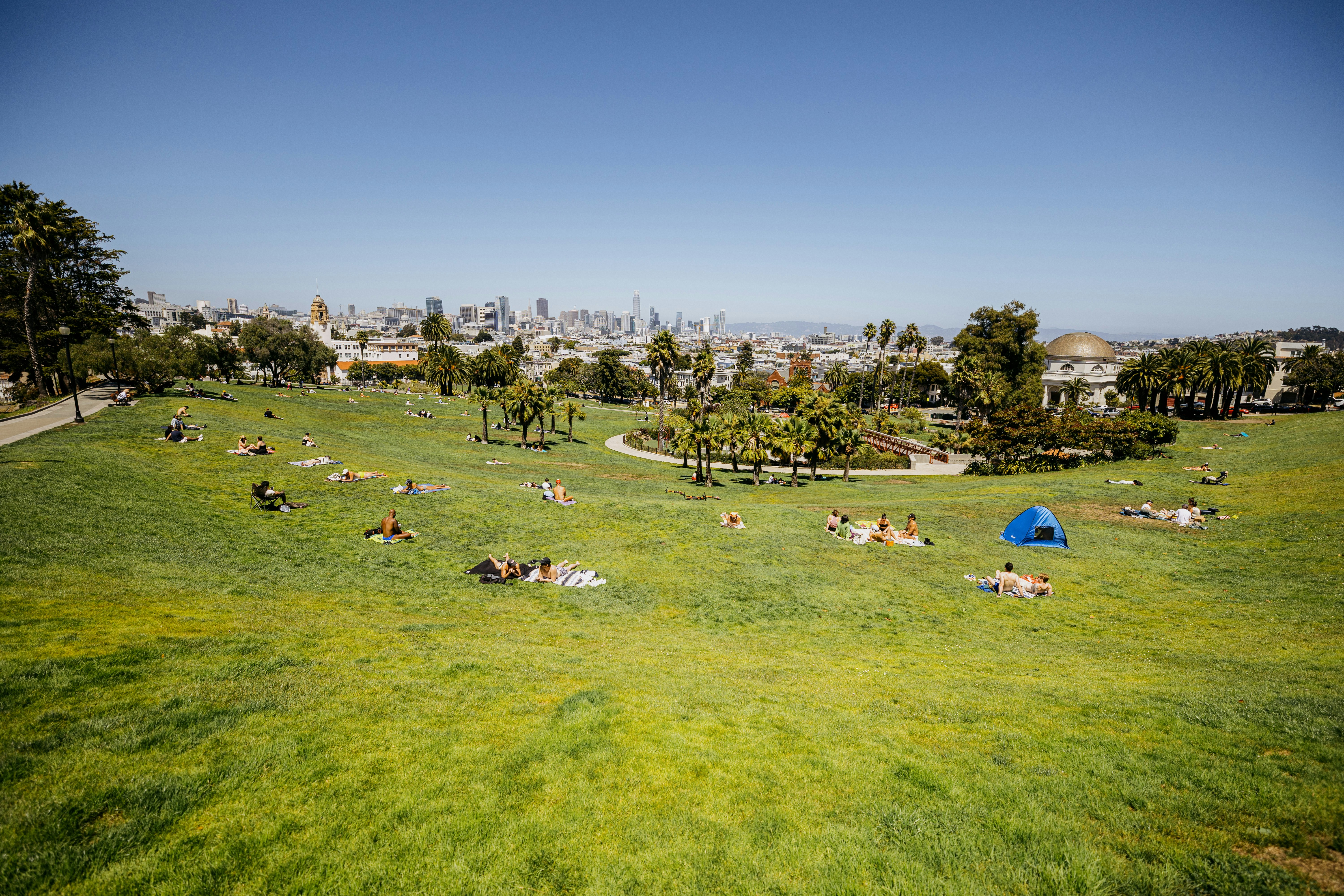 Green park with people relaxing, blue tent, and distant city.