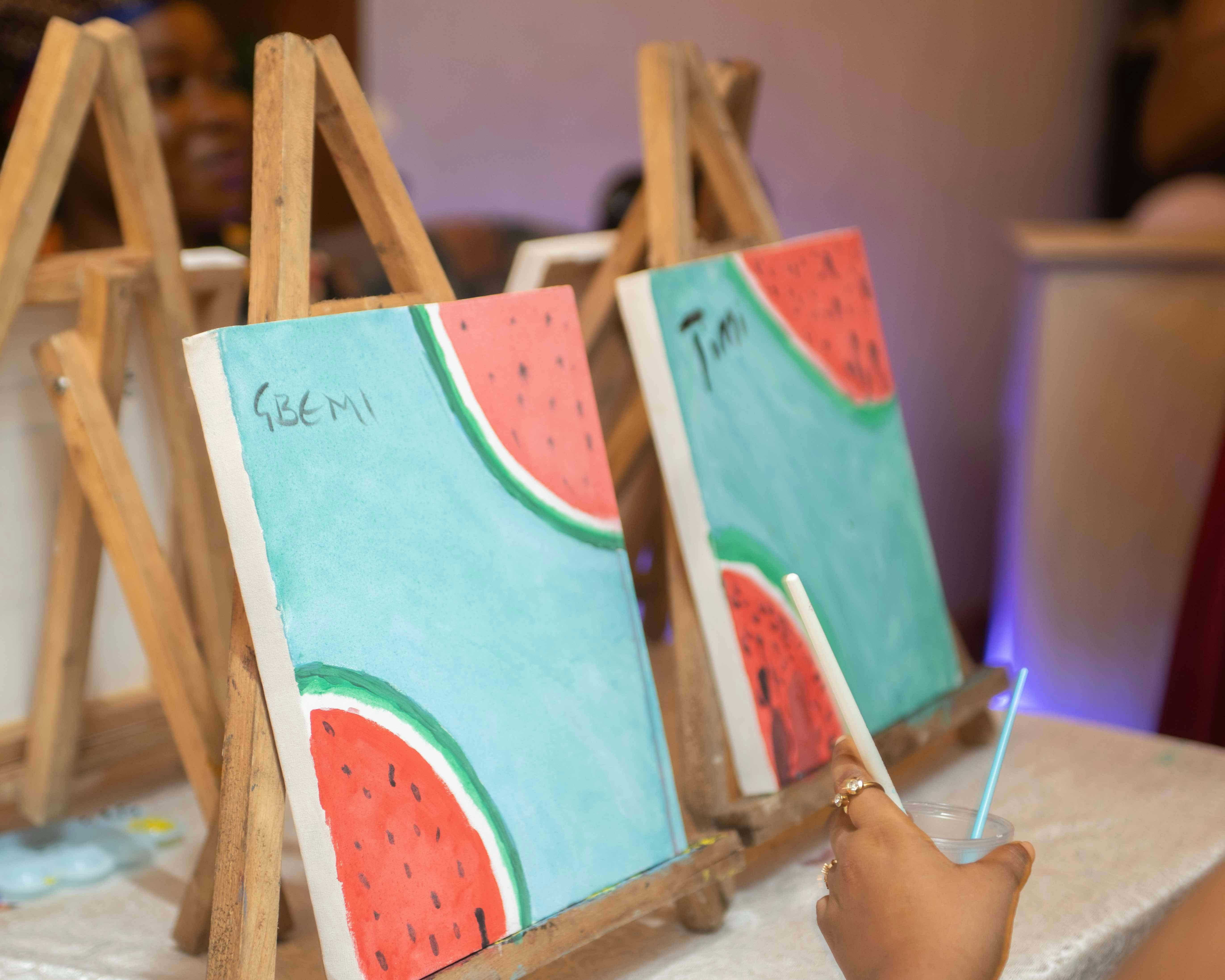 Watermelon paintings on easels, with a hand holding a paintbrush.
