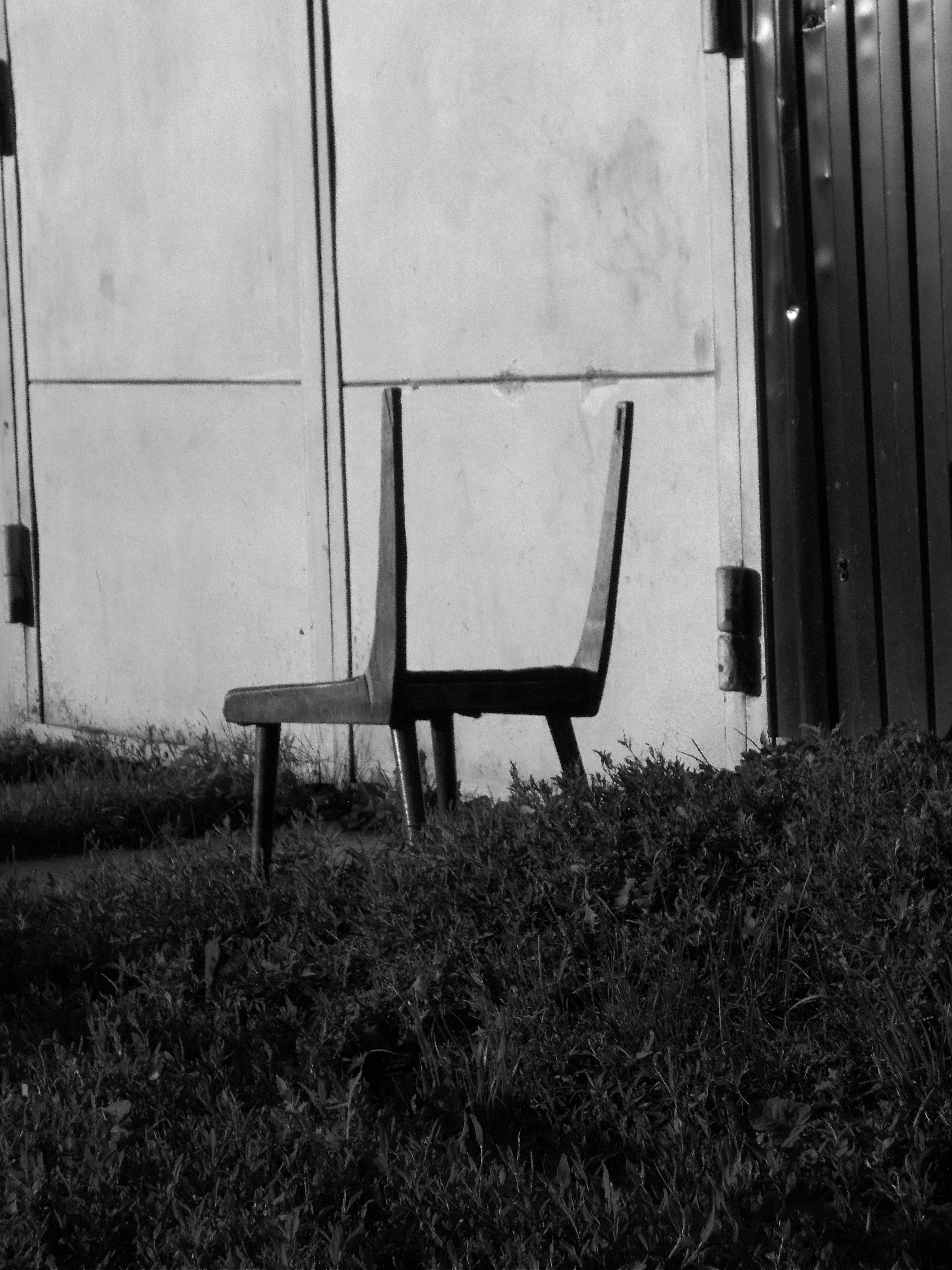 A solitary chair sits in front of a weathered wall and door, casting a long shadow over tall grass in black-and-white tones. This image emphasizes solitude and decay without color.