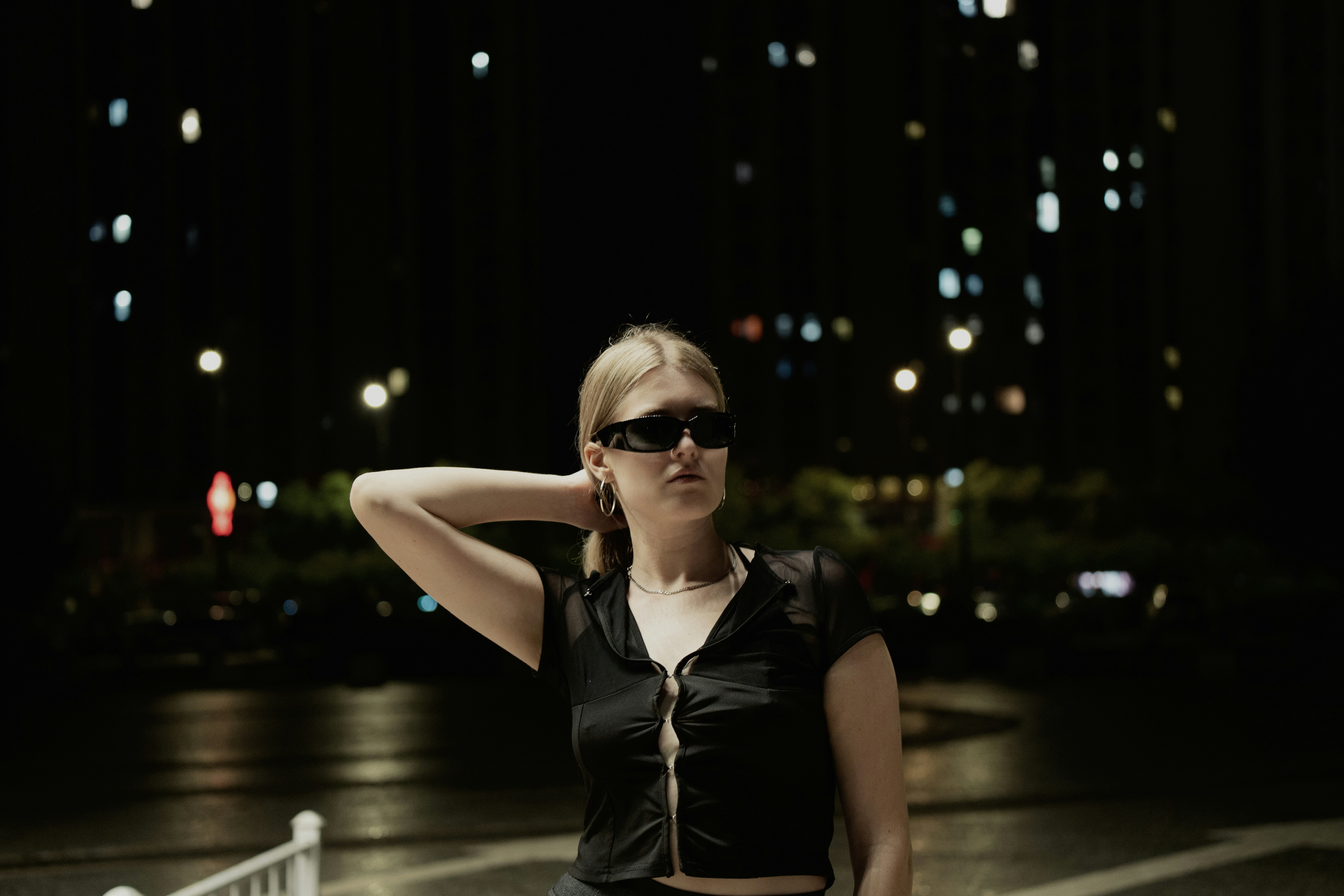 Woman wearing sunglasses poses at night with city lights.