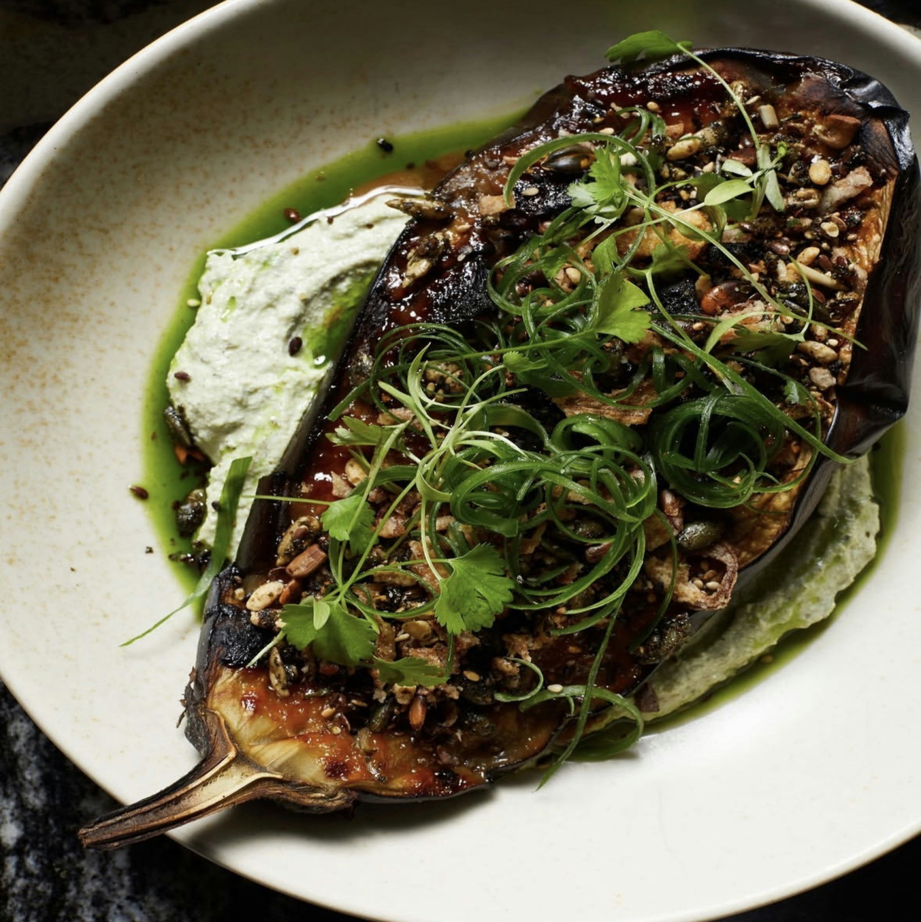 Charred miso-glazed eggplant half on pale green herb tahini with curled scallions, cilantro, herb oil, and toasted seed crunch