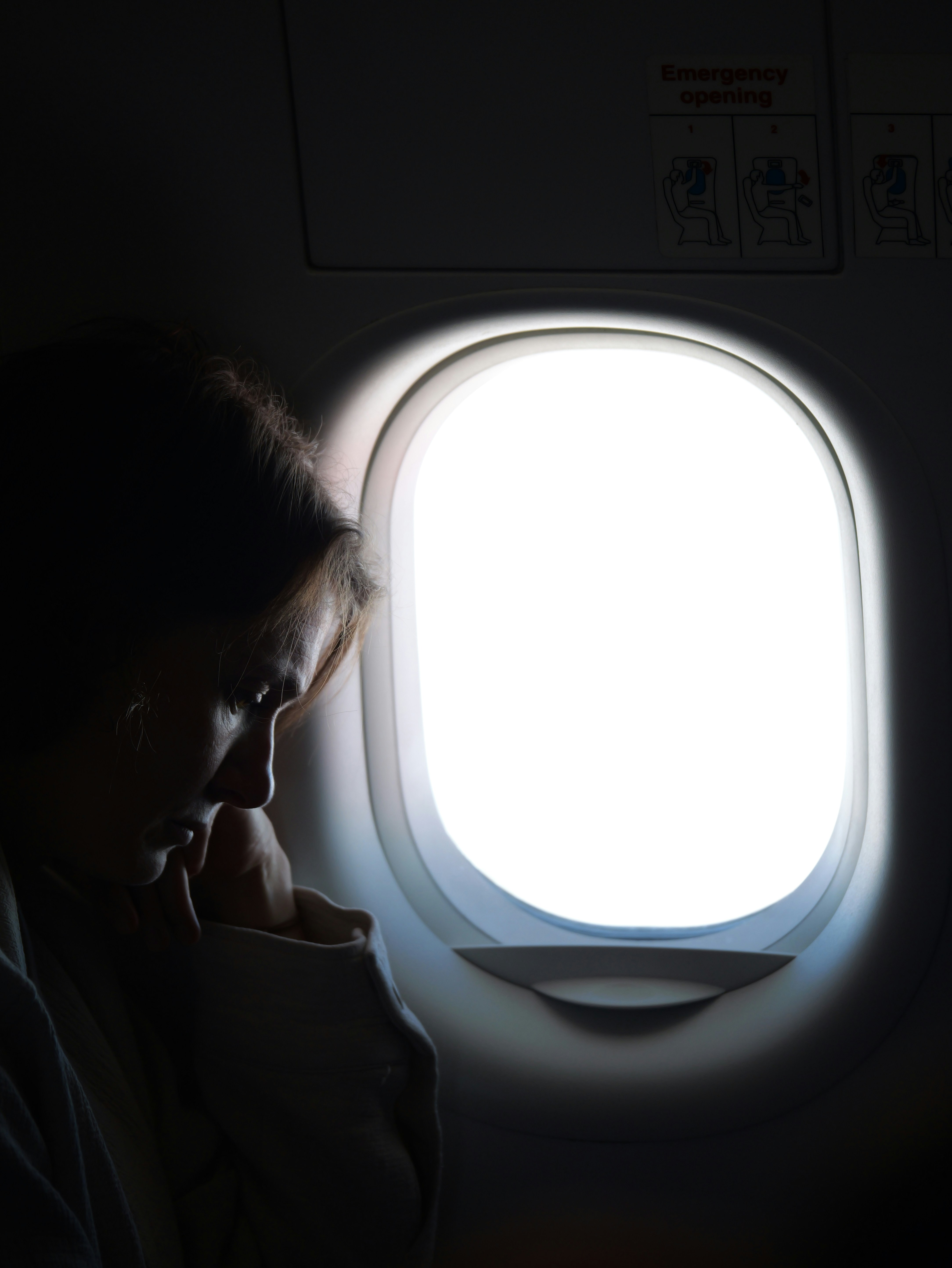 Woman sits by bright airplane window in dark cabin.