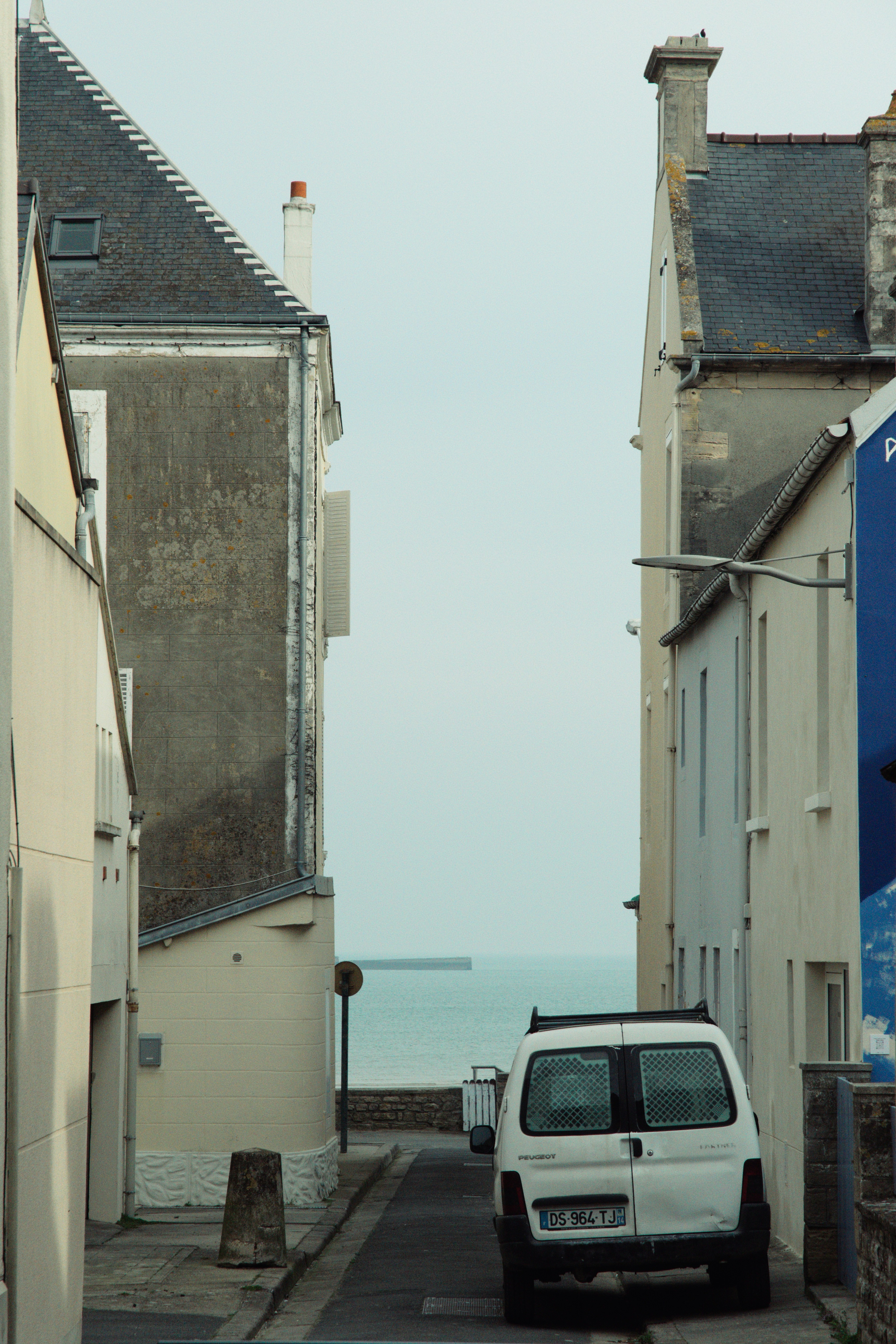 Narrow empty coastal village street between stone houses opening onto a calm sea under soft grey light
