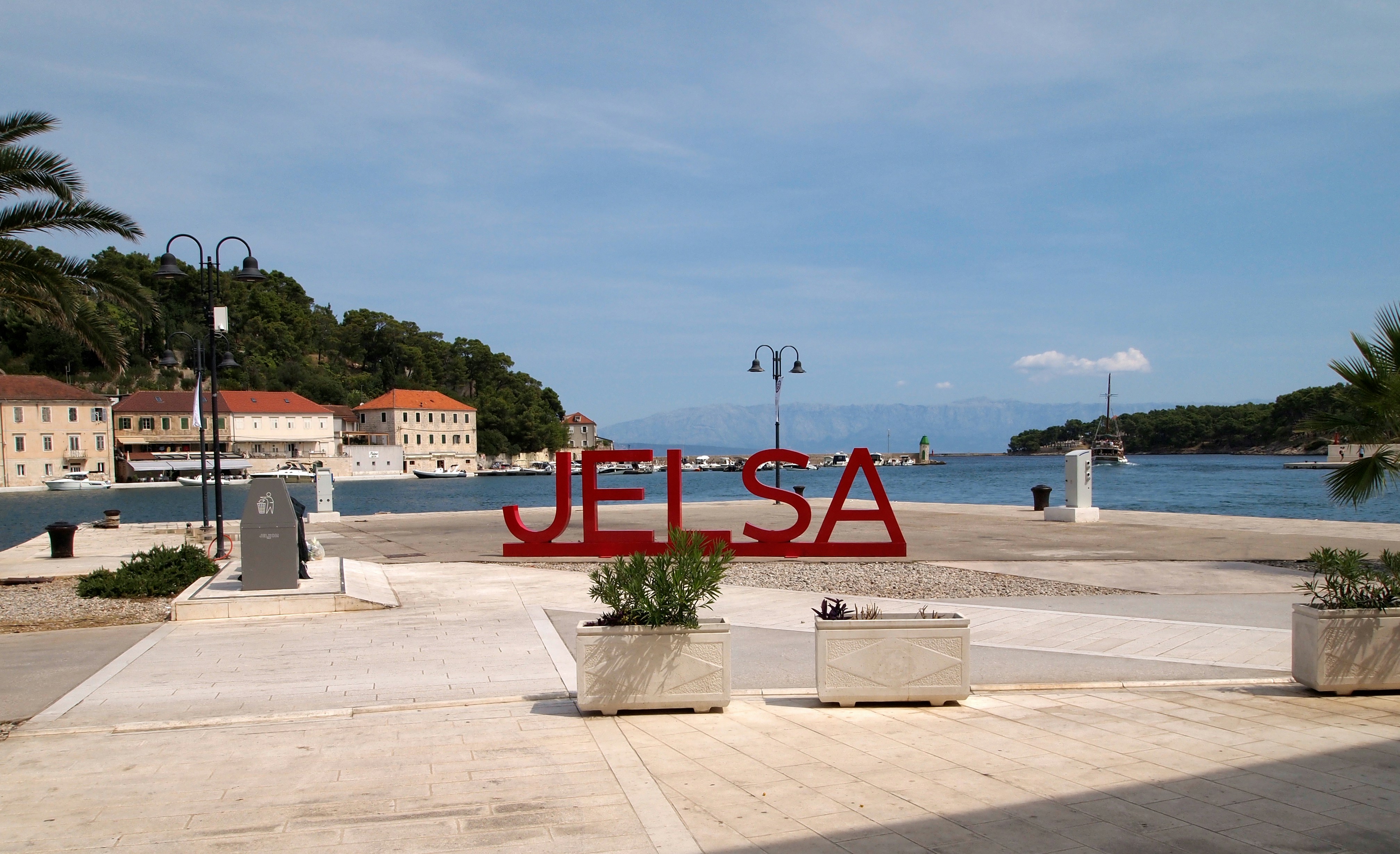 Insegna rossa “JELSA” sul lungomare con barche in mare e spiaggia rocciosa sullo sfondo, Hvar.