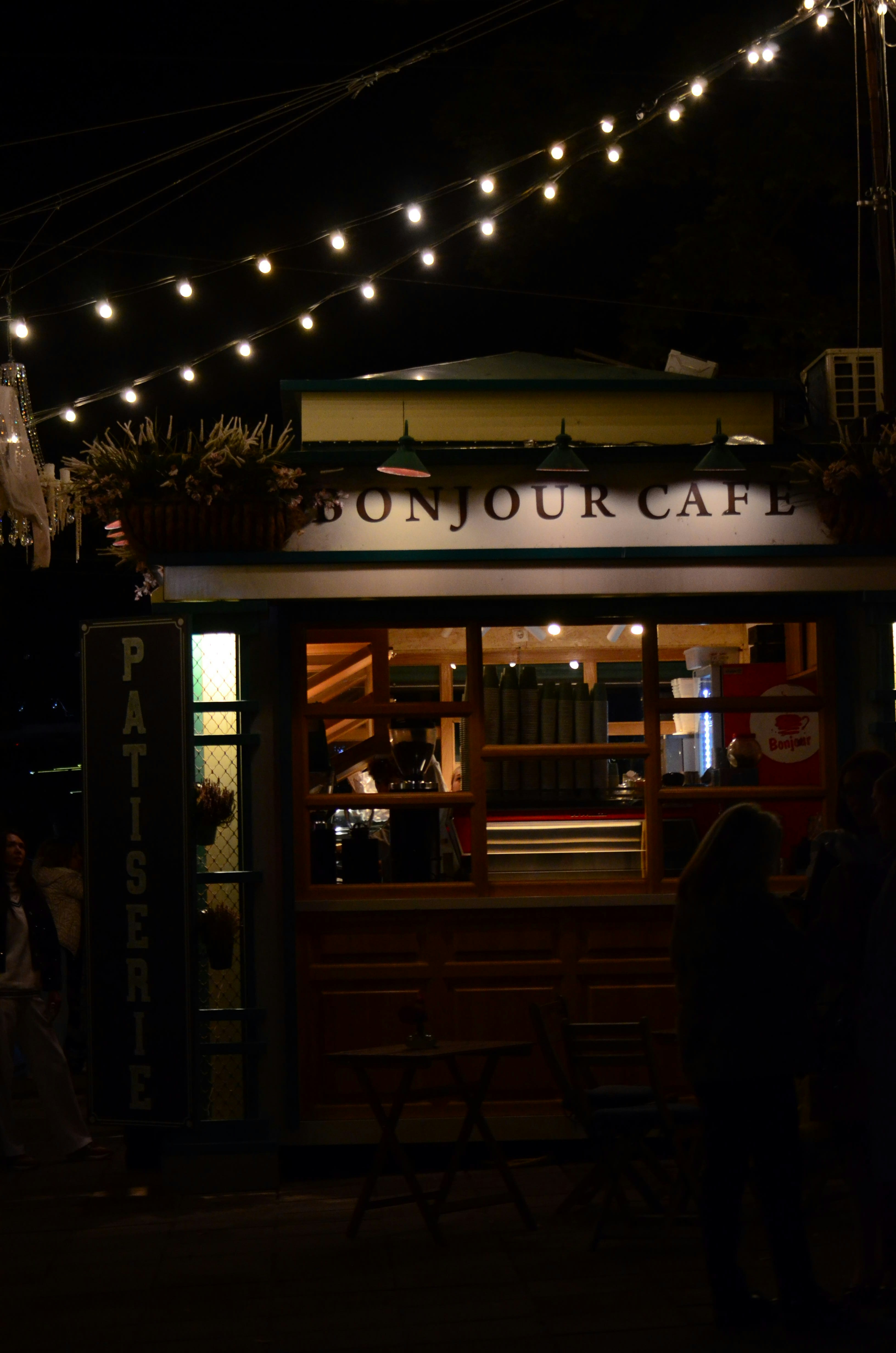 A small cafe kiosk labeled "BONJOUR CAFE" and "PATISSERIE" under string lights at night