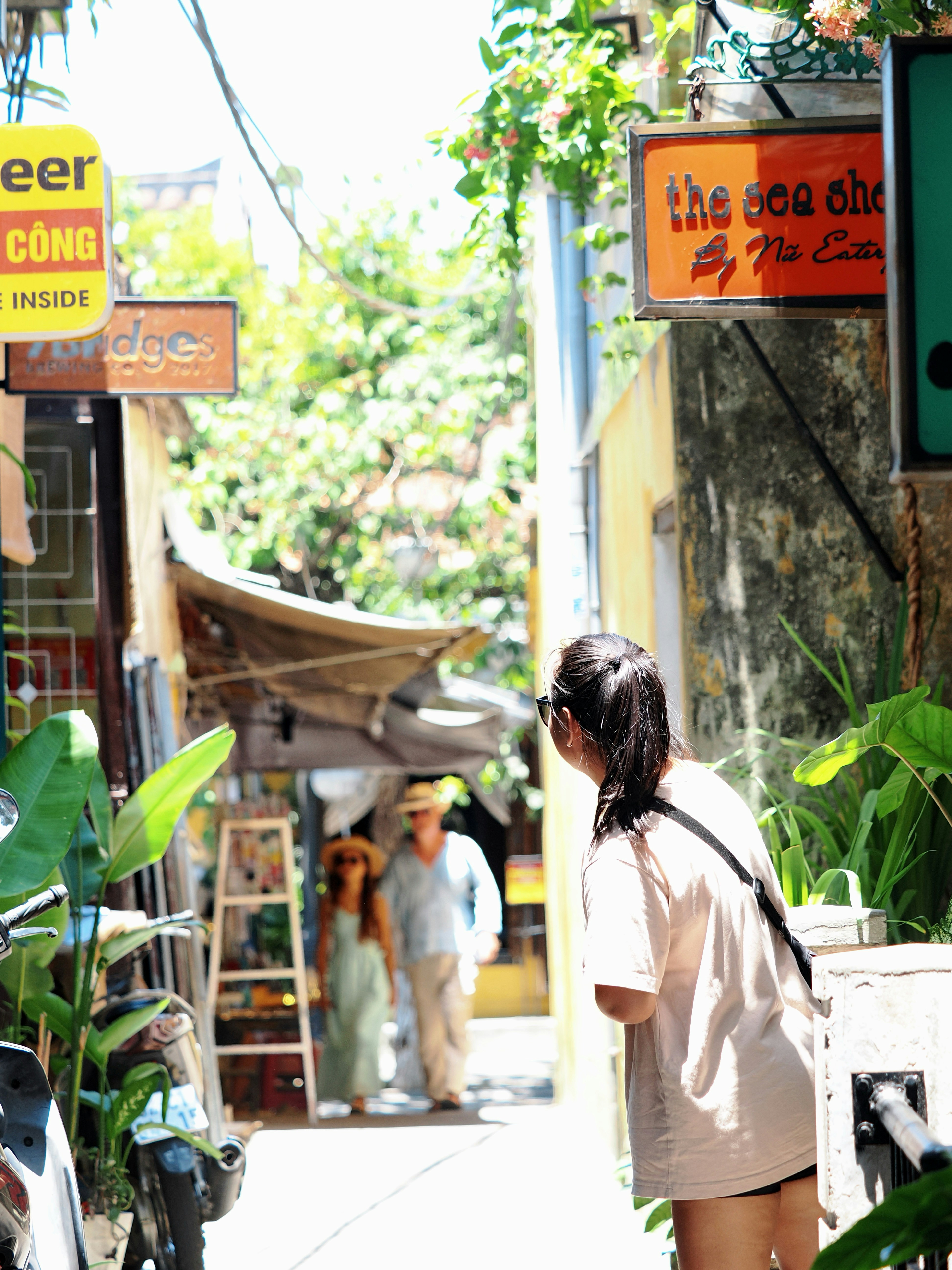 Woman looking down a sunny, narrow alleyway with shops.