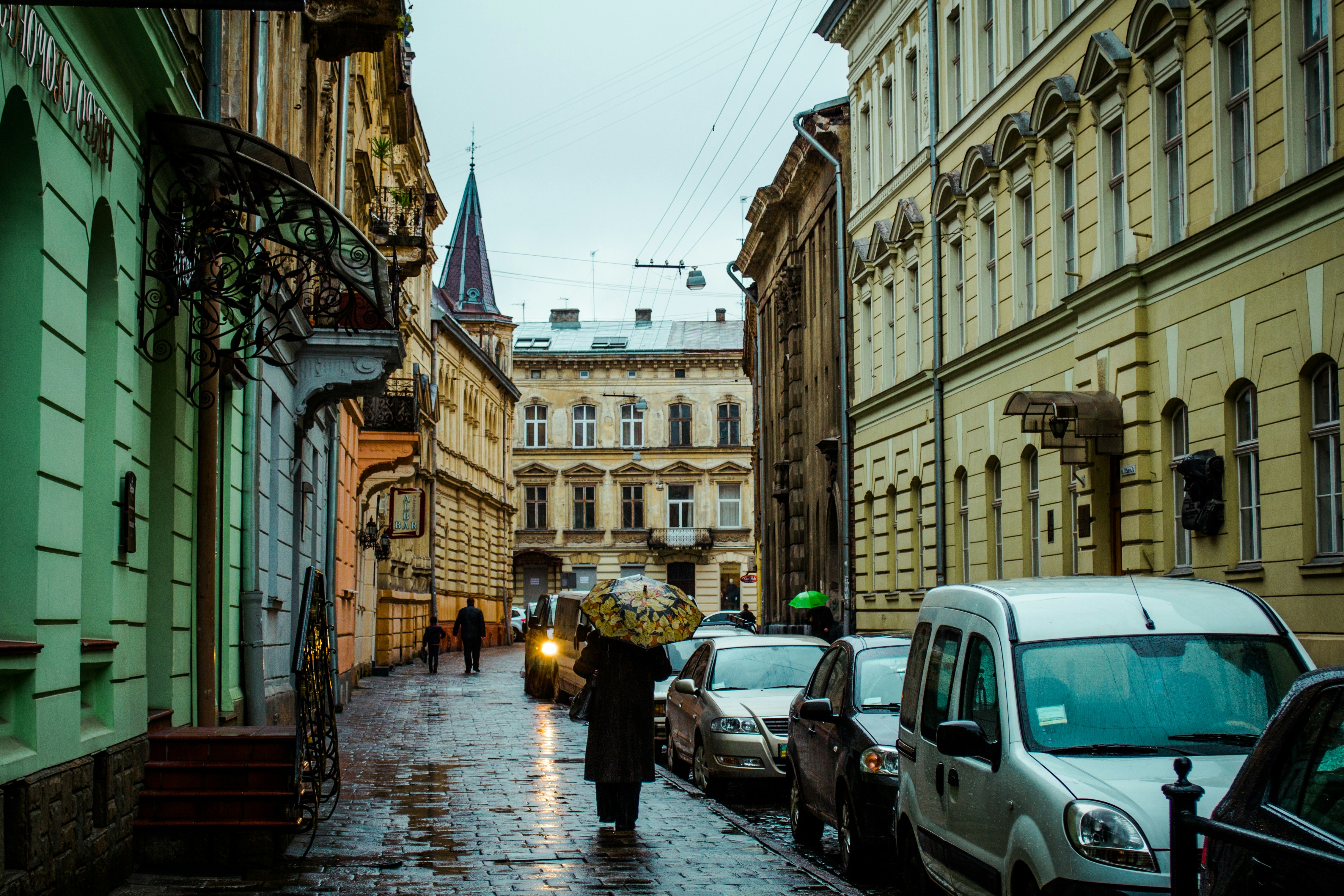 Beautiful cinematic street photography capturing a cozy rainy day in the old town of Lviv. The image showcases vintage multi-story architecture with ornate facades, a distant spire roof, and pedestrians with colorful umbrellas walking down a wet, glossy cobblestone road that reflects the soft ambient light, flanked by parked vehicles.