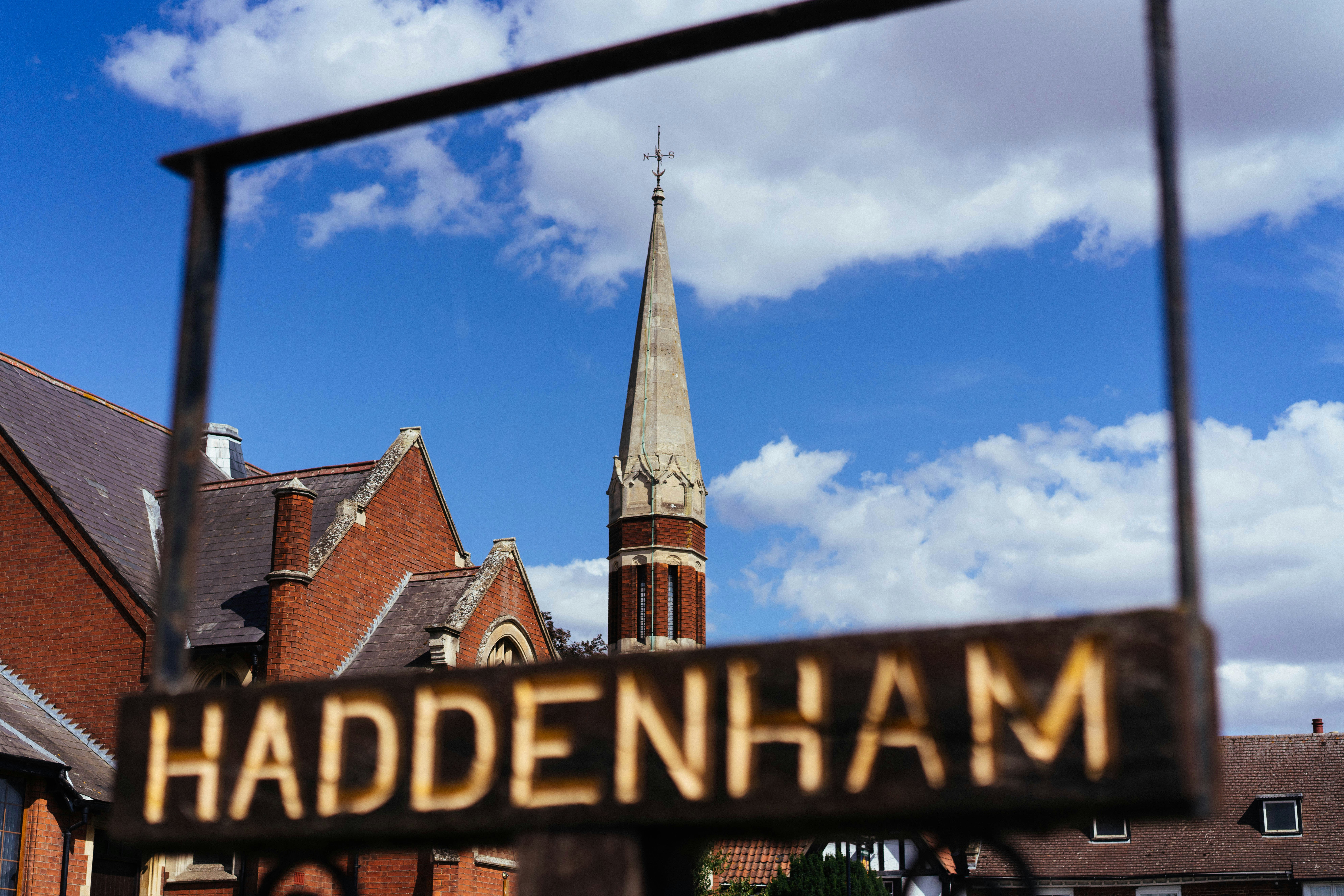 Blurred haddenham sign frames a church steeple under blue sky.