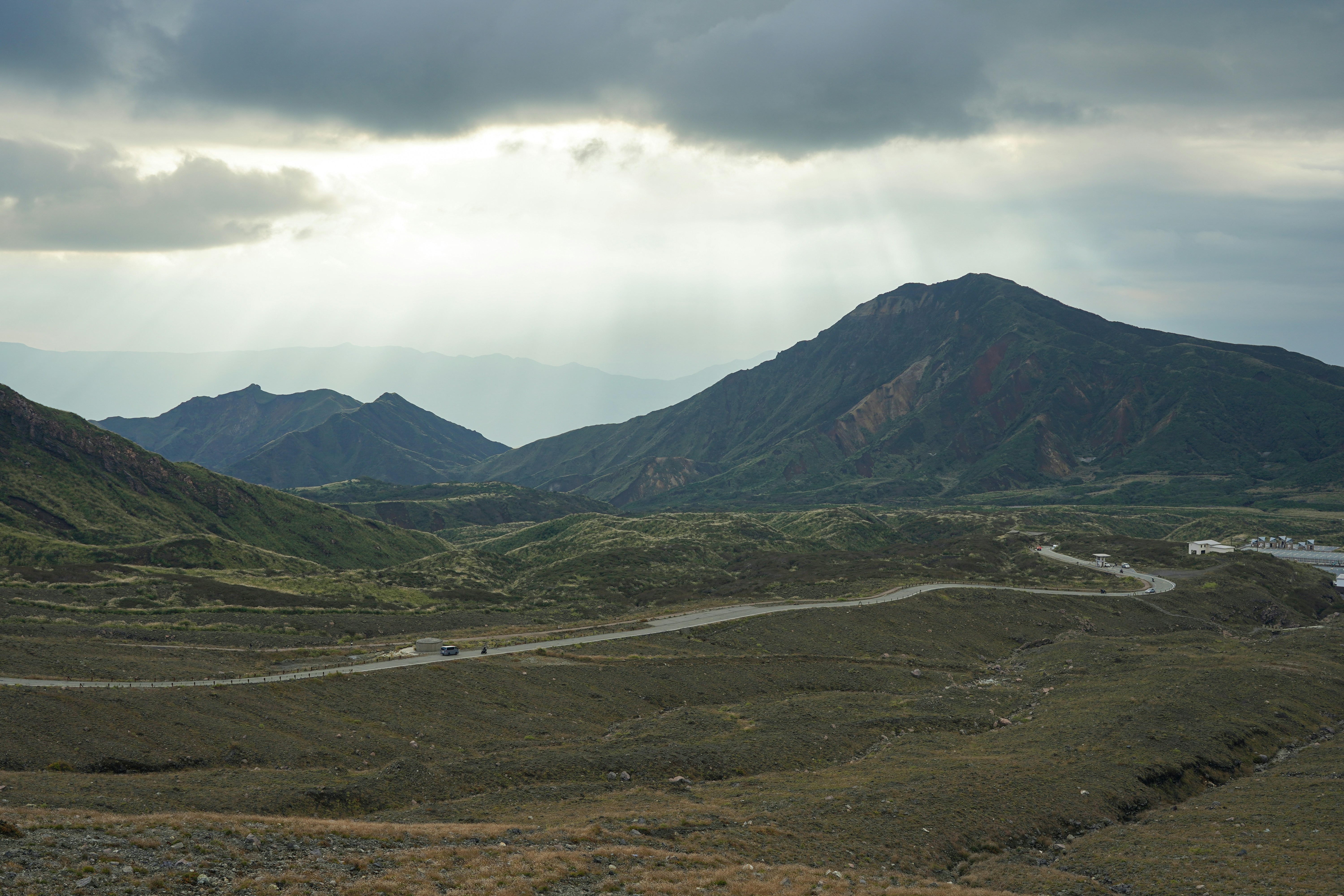 A road winds across the grassy upper slopes of Aso as sunbeams break through storm cloud over the volcanic peaks
