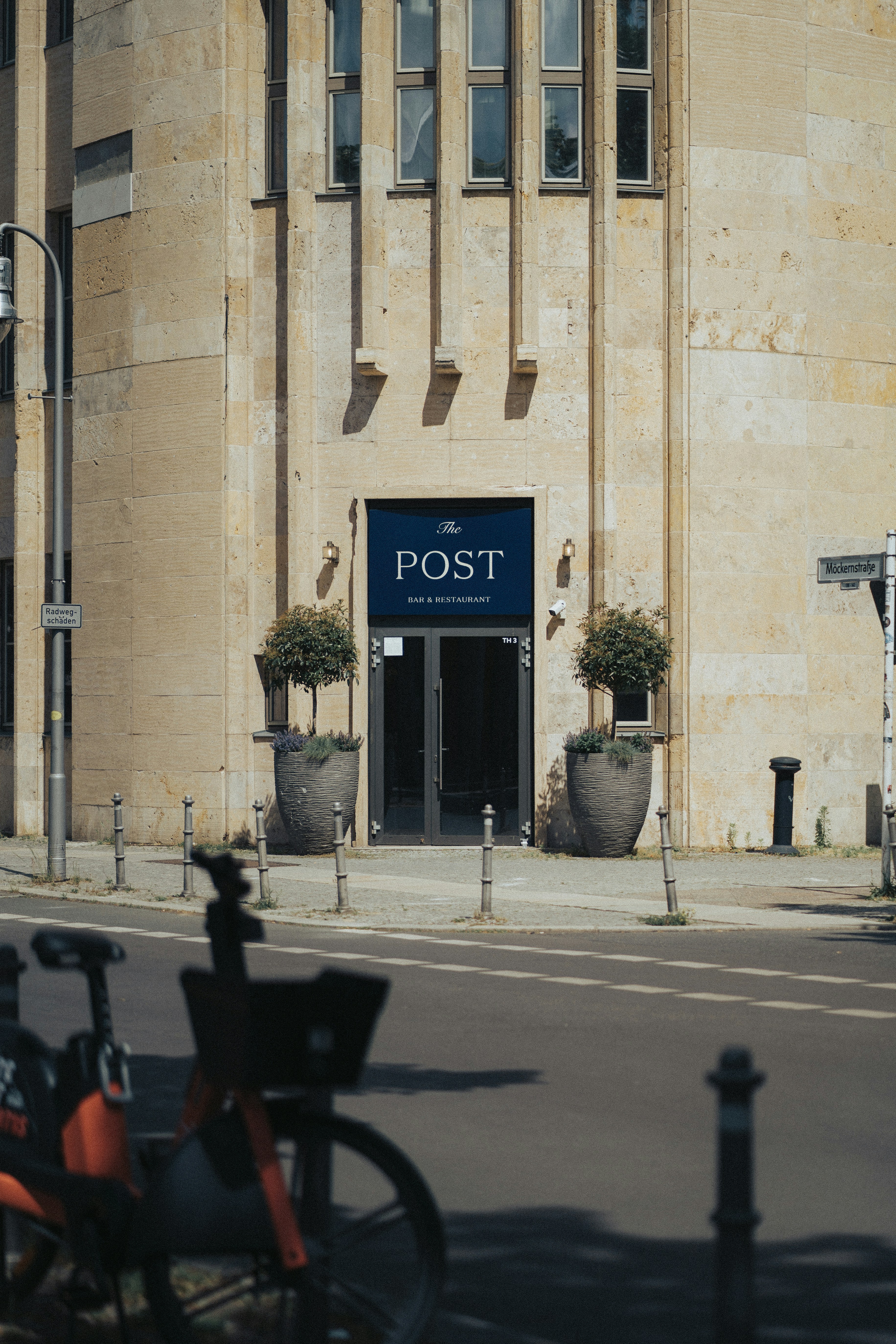 Eingang zum steinernen Gebäude mit "Pfosten"-Schild und Topfbäumen.