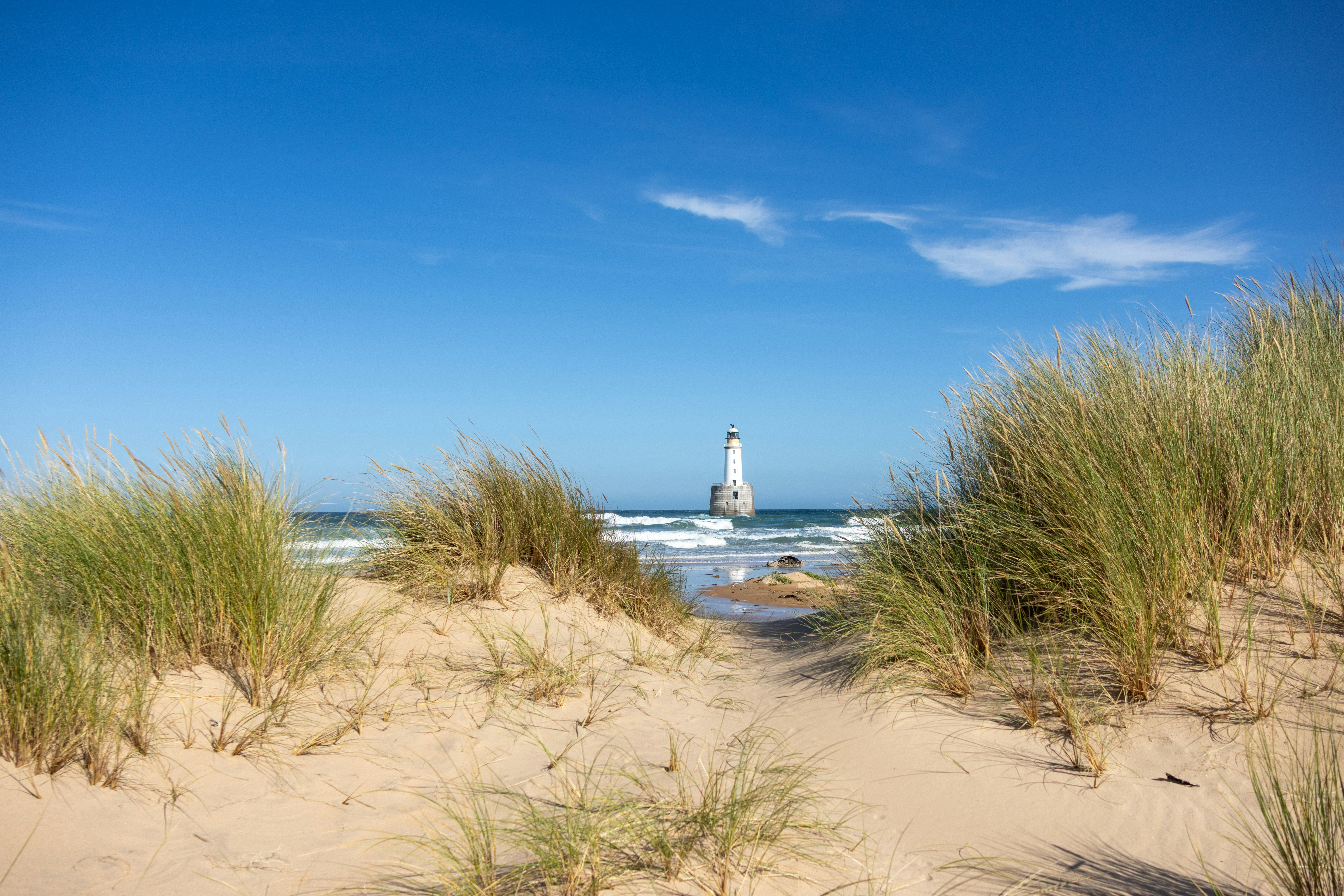 Sandy beach path leading to a lighthouse under blue sky.
