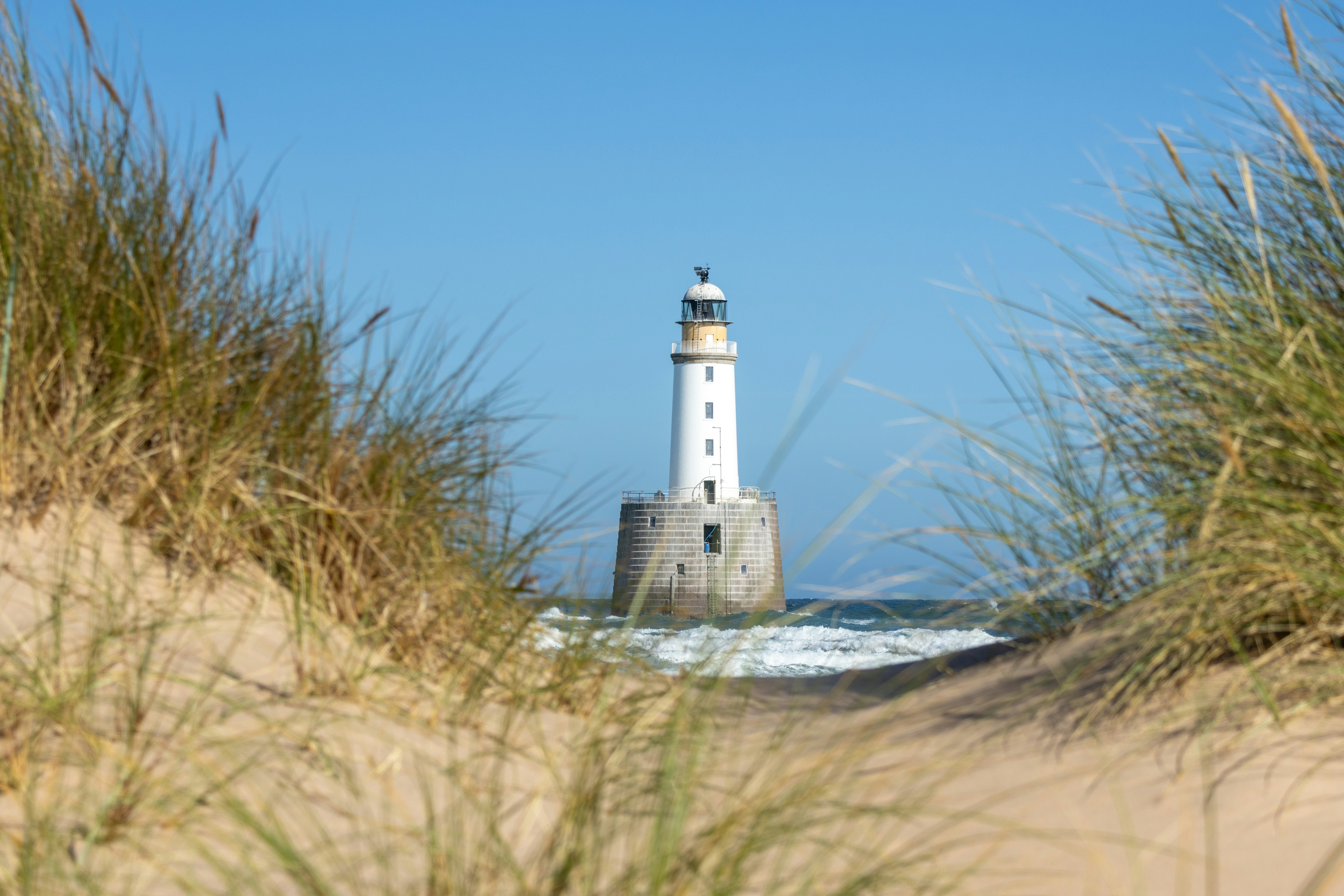 Lighthouse in the ocean, viewed through grassy sand dunes