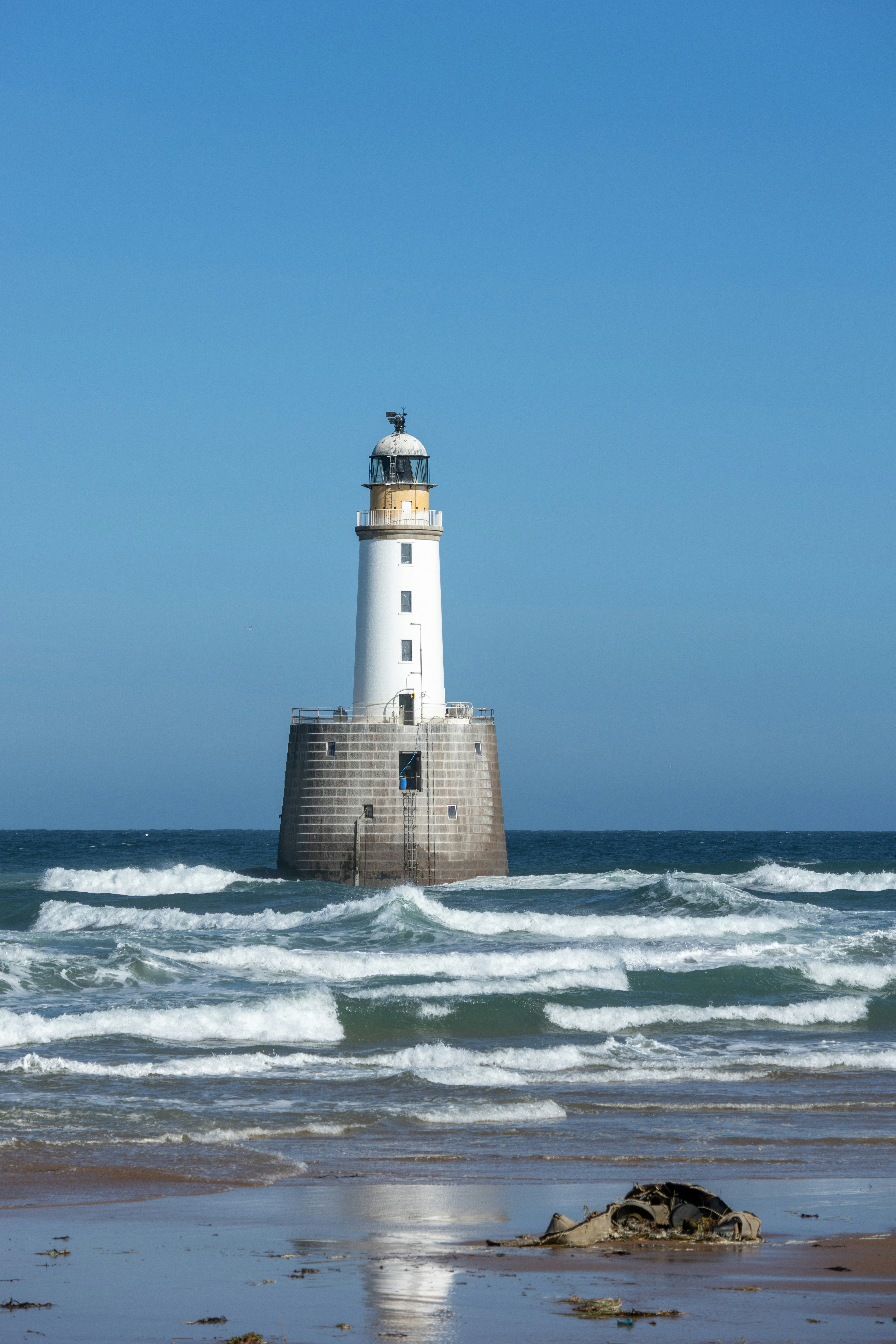 White lighthouse stands amidst ocean waves under a blue sky.