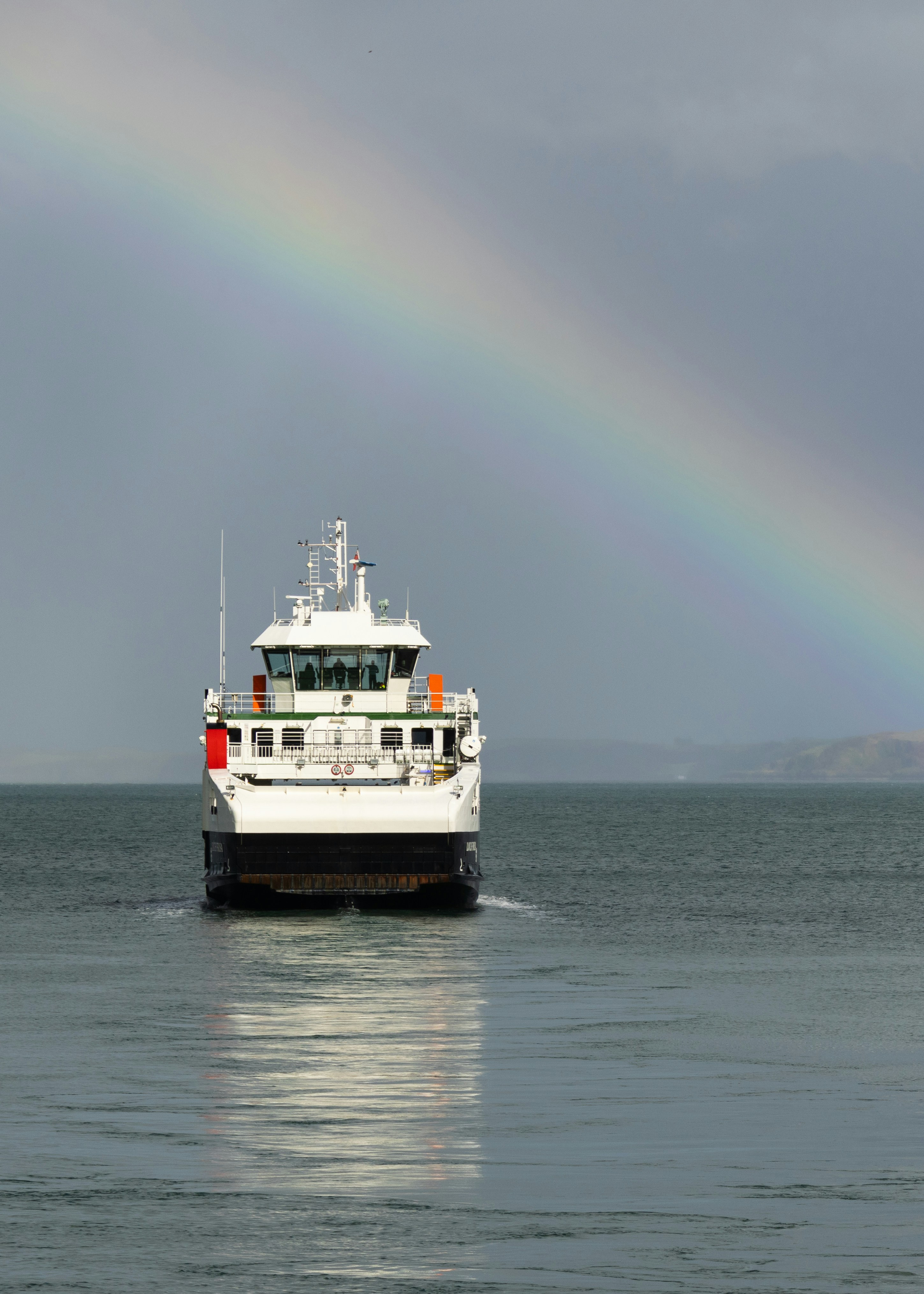 A white ferry boat sailing under a vibrant rainbow.