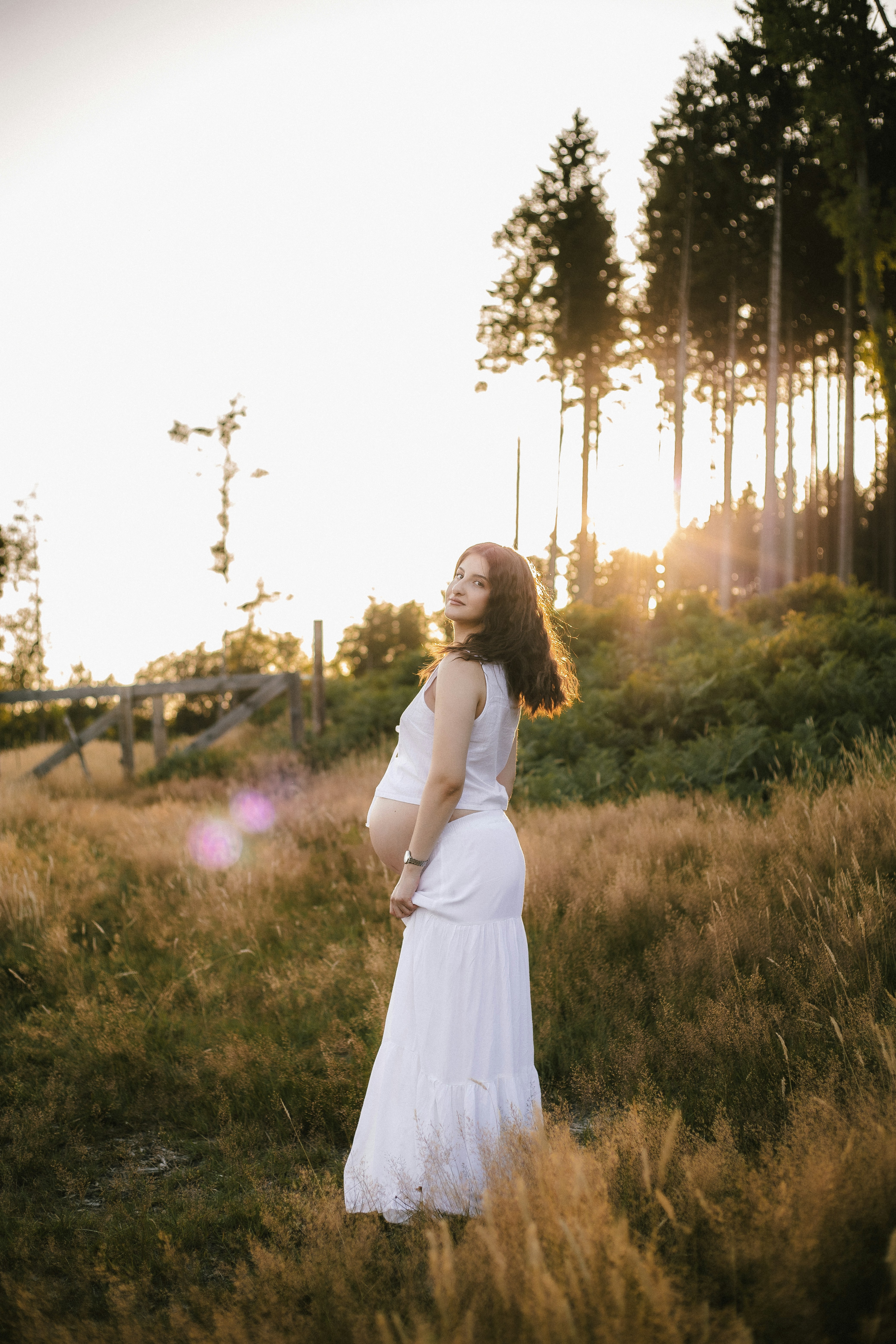 A pregnant woman stands in a field at sunset.