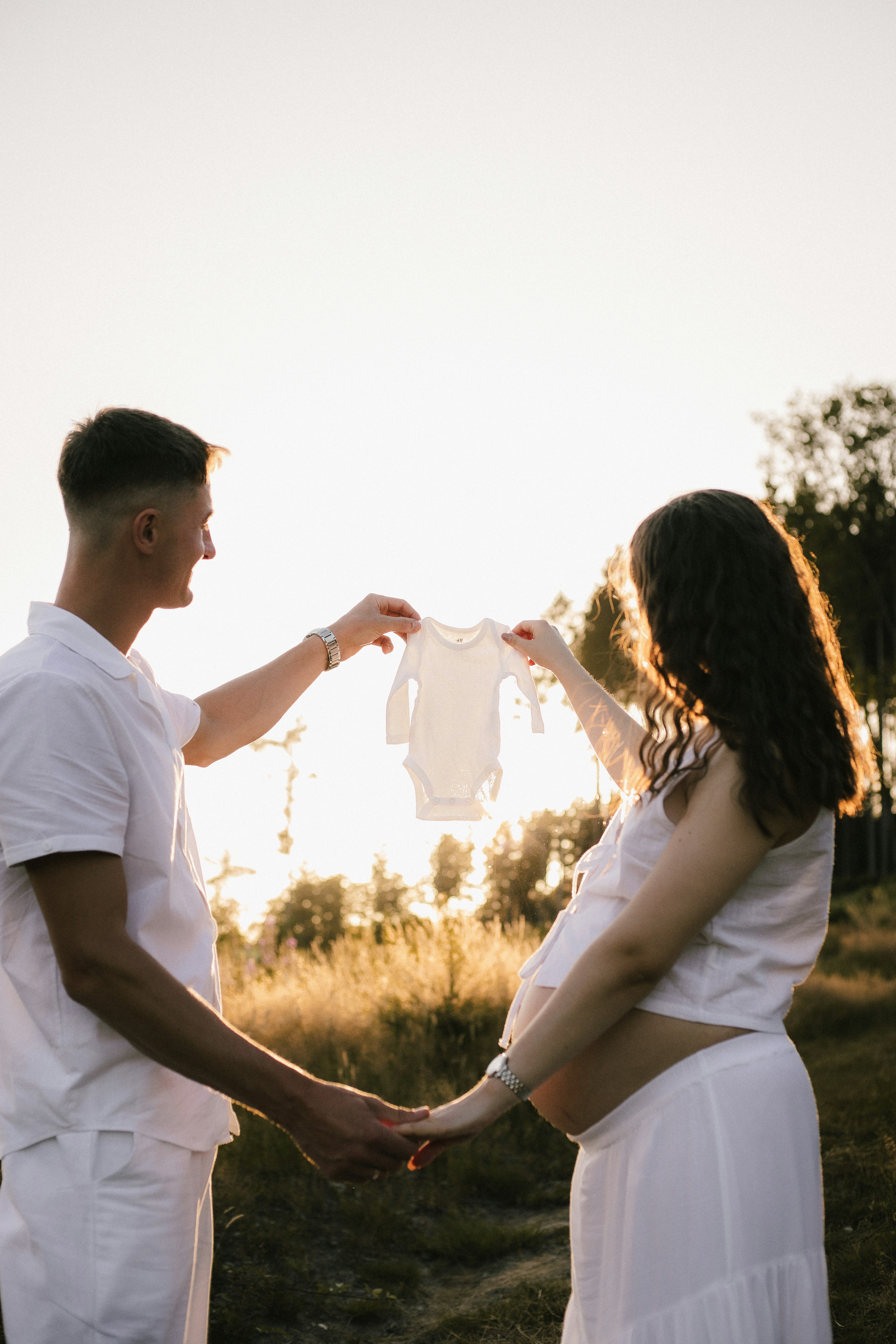 Pregnant couple holding baby onesie at sunset.