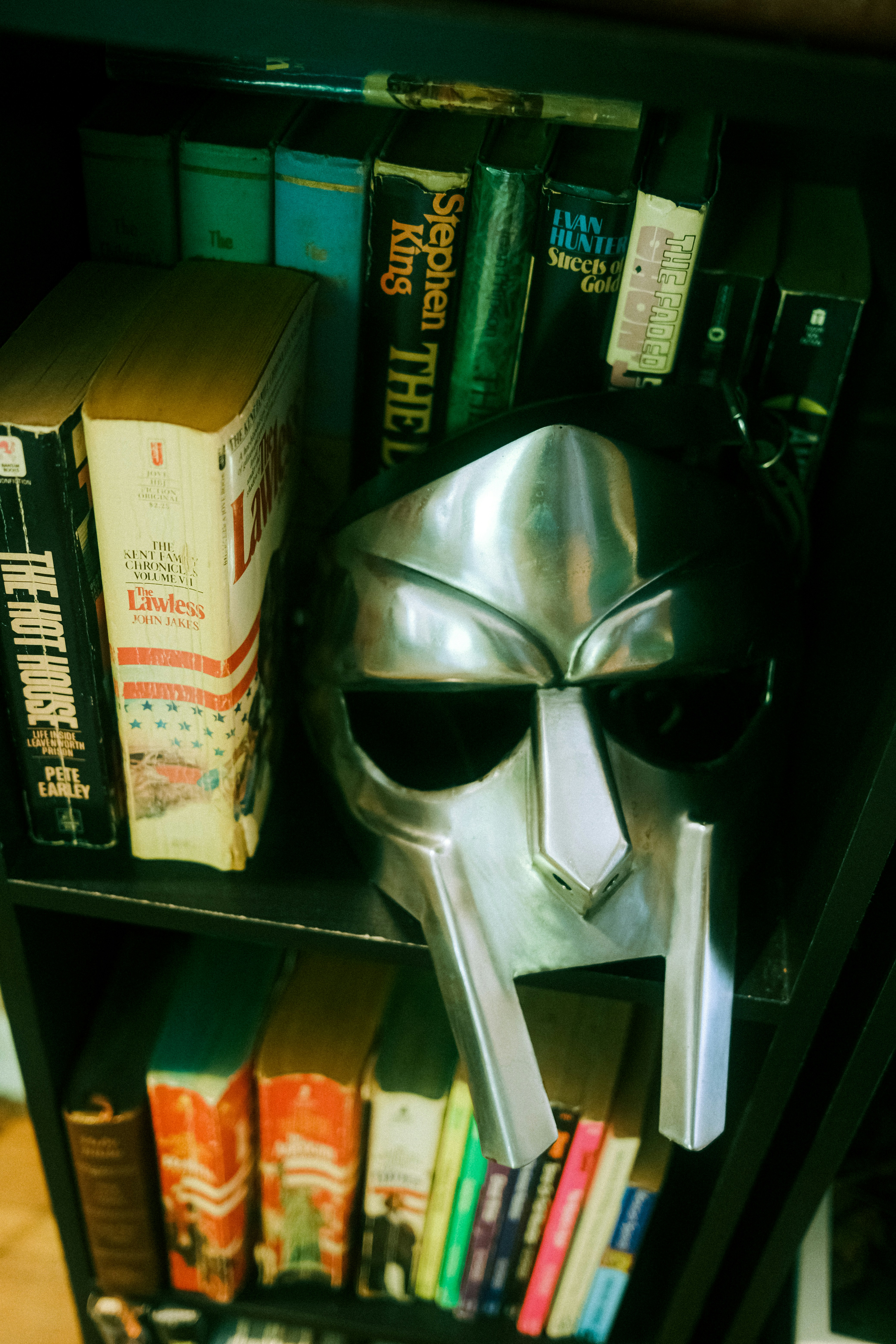 Metal mask displayed on a dark bookshelf among various books.