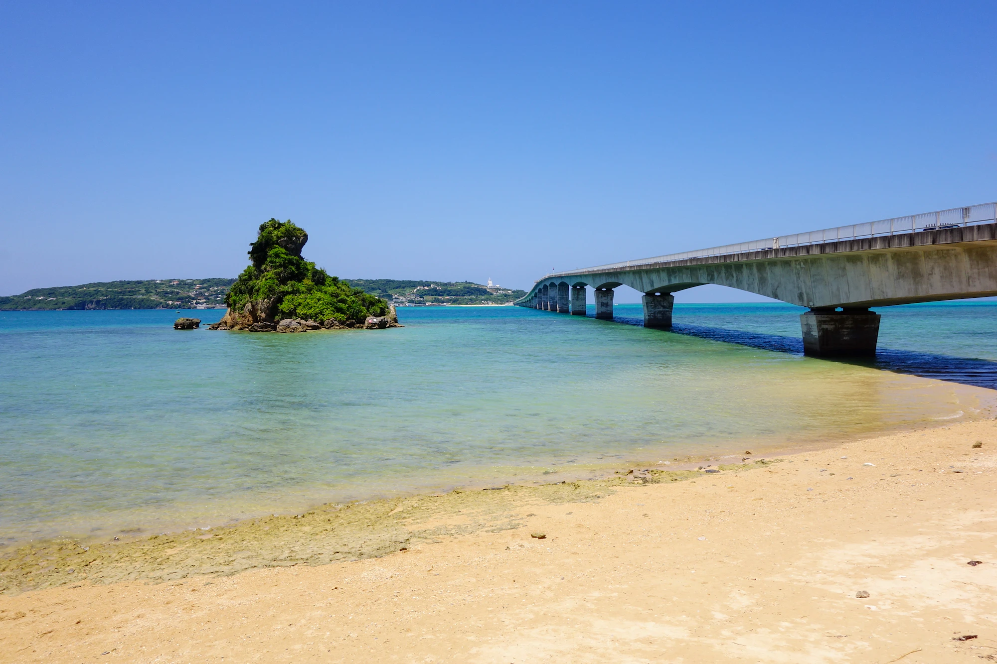 A white sand beach borders clear turquoise water beside a small green island and a low bridge.