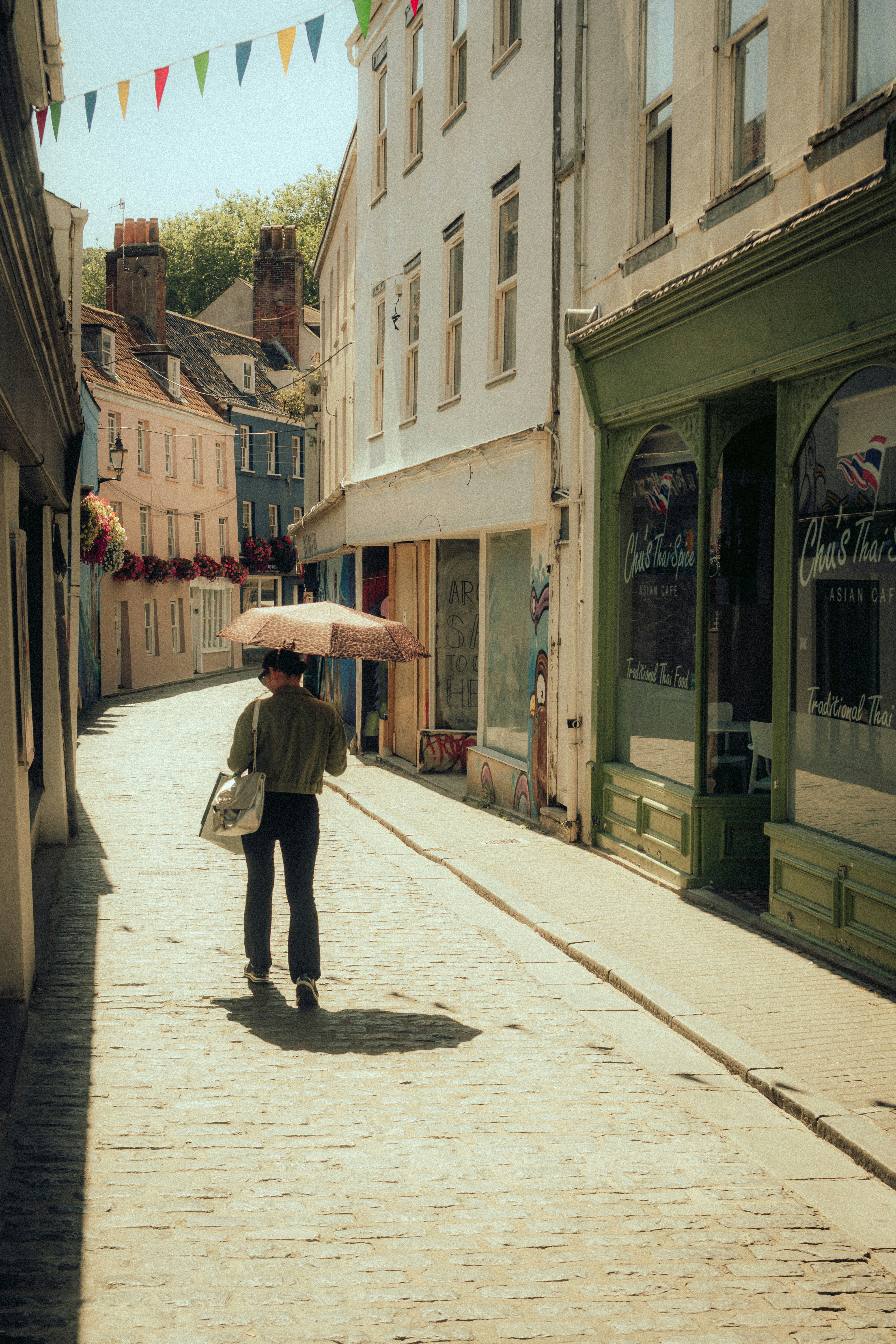 Person walks narrow street under colorful flags holding umbrella.
