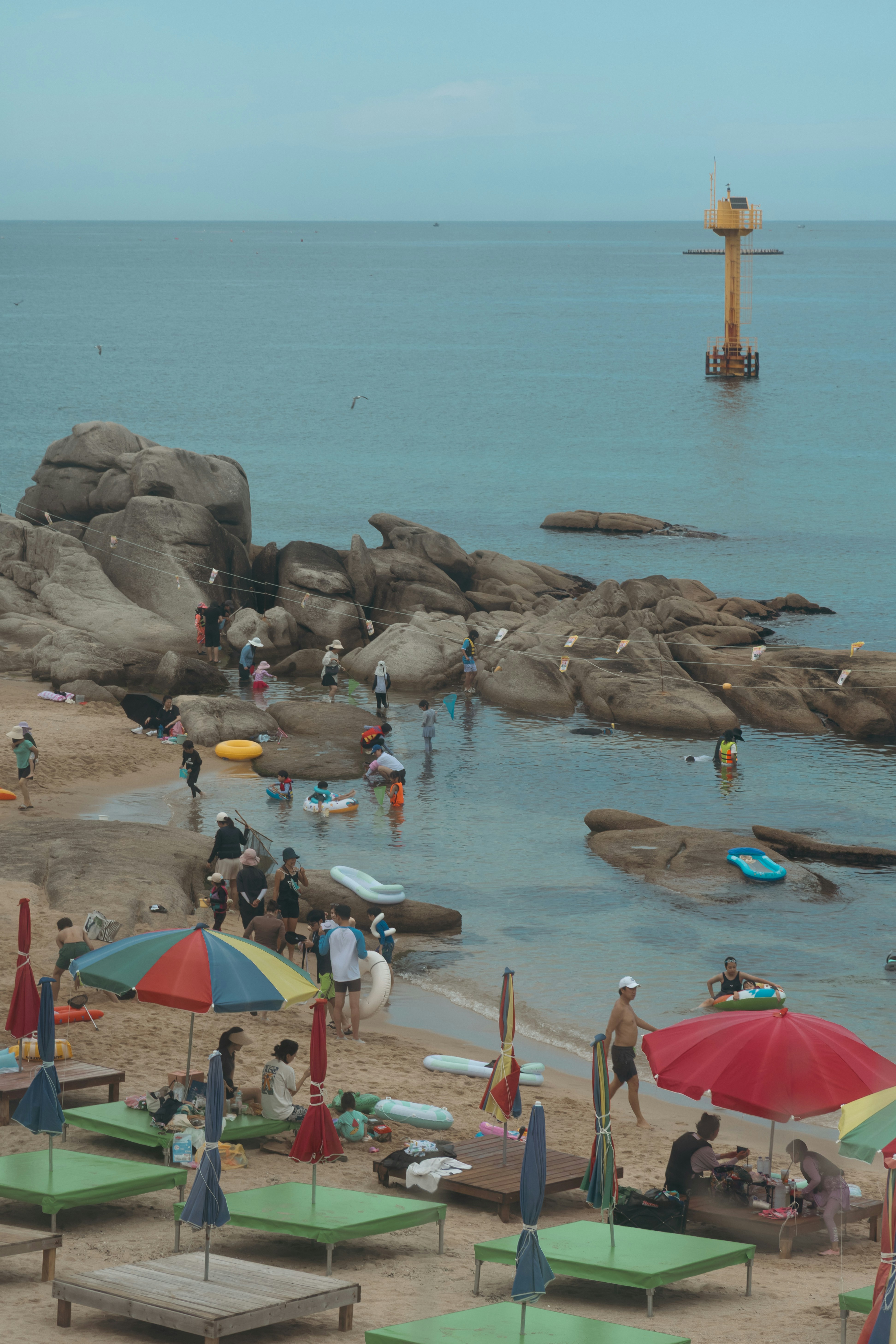 Crowded beach with people, rocks, colorful umbrellas, and ocean buoy