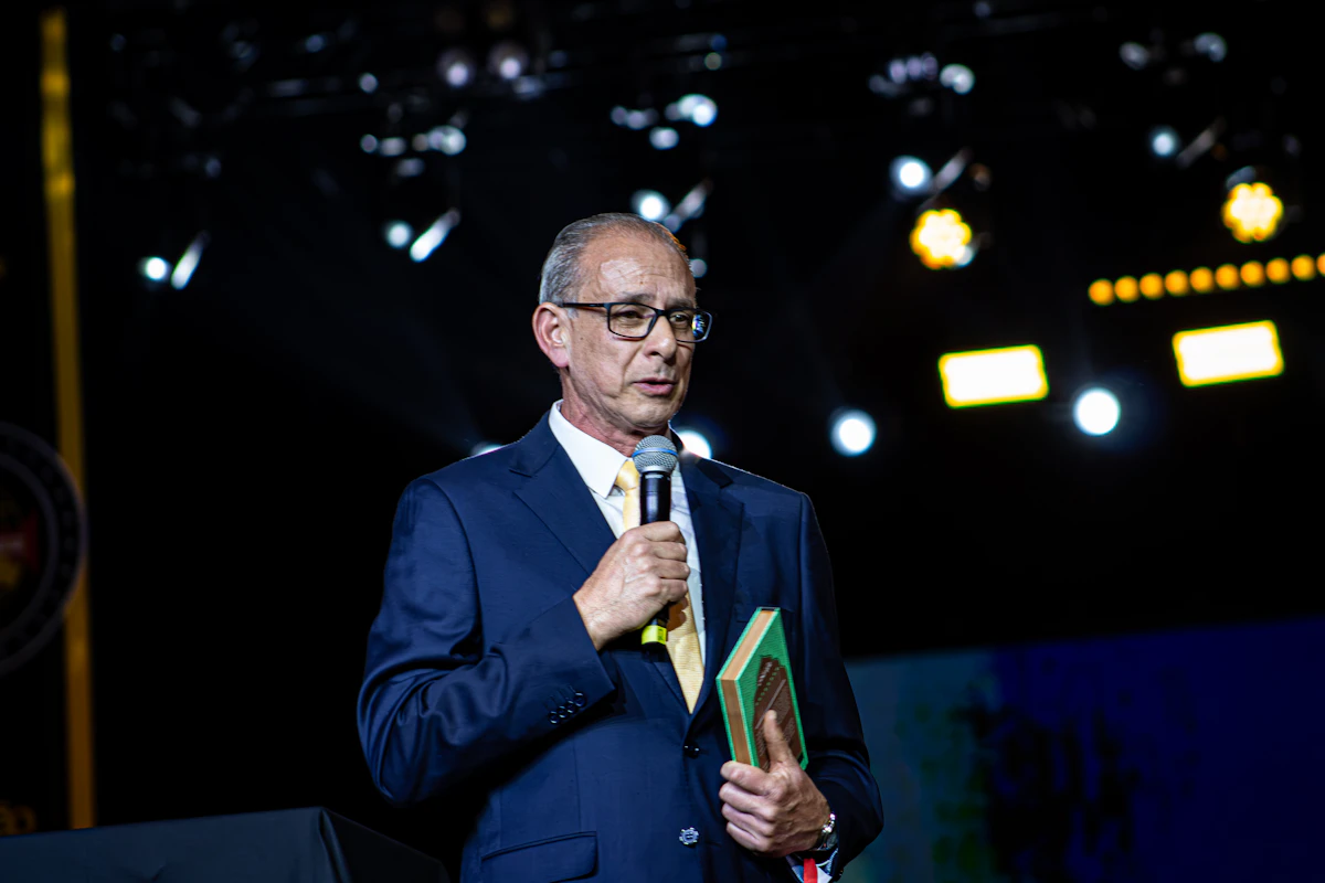 An author in a suit speaks into a microphone on stage while holding a copy of a book, promoting it to an audience