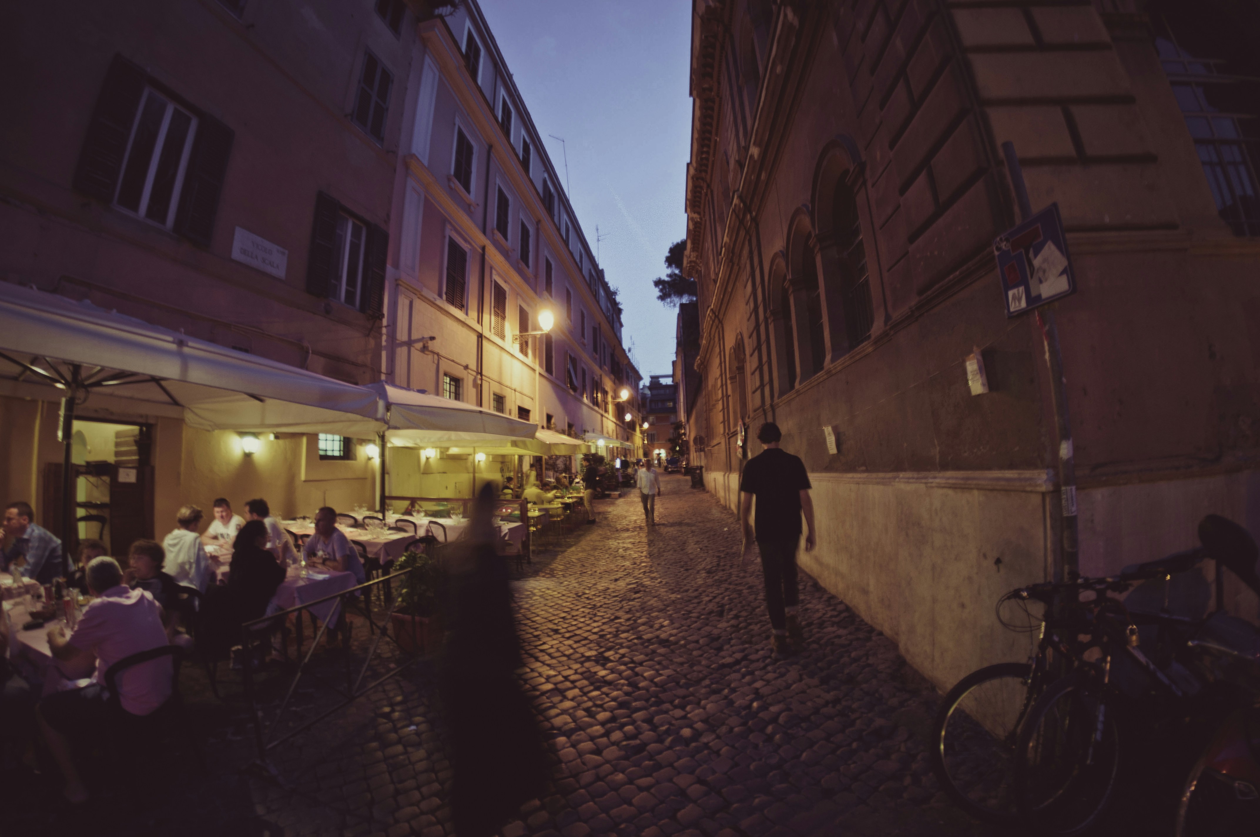 People dining at tables in a narrow stone street at dusk