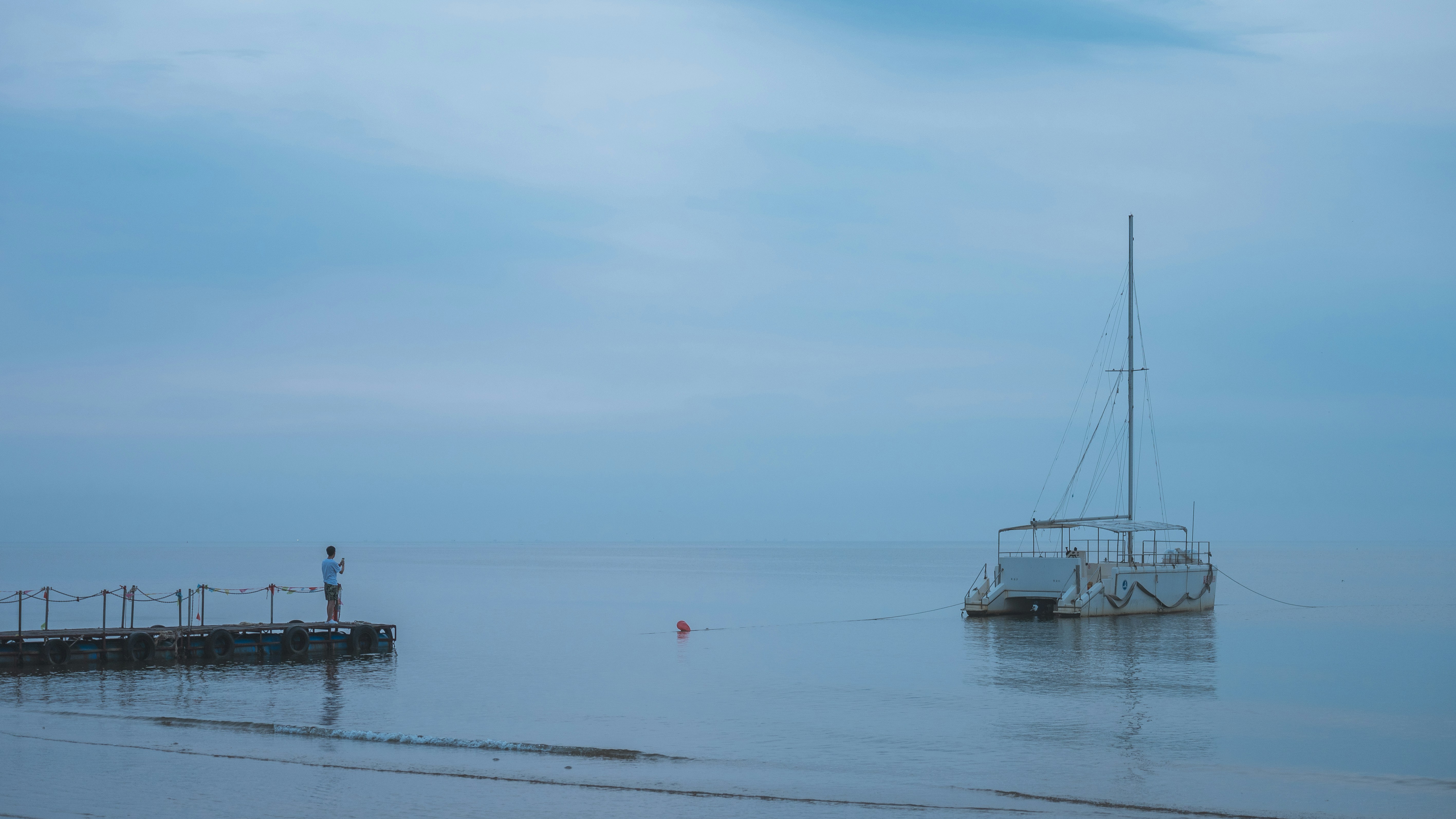 Person on pier near sailboat in calm, misty water.