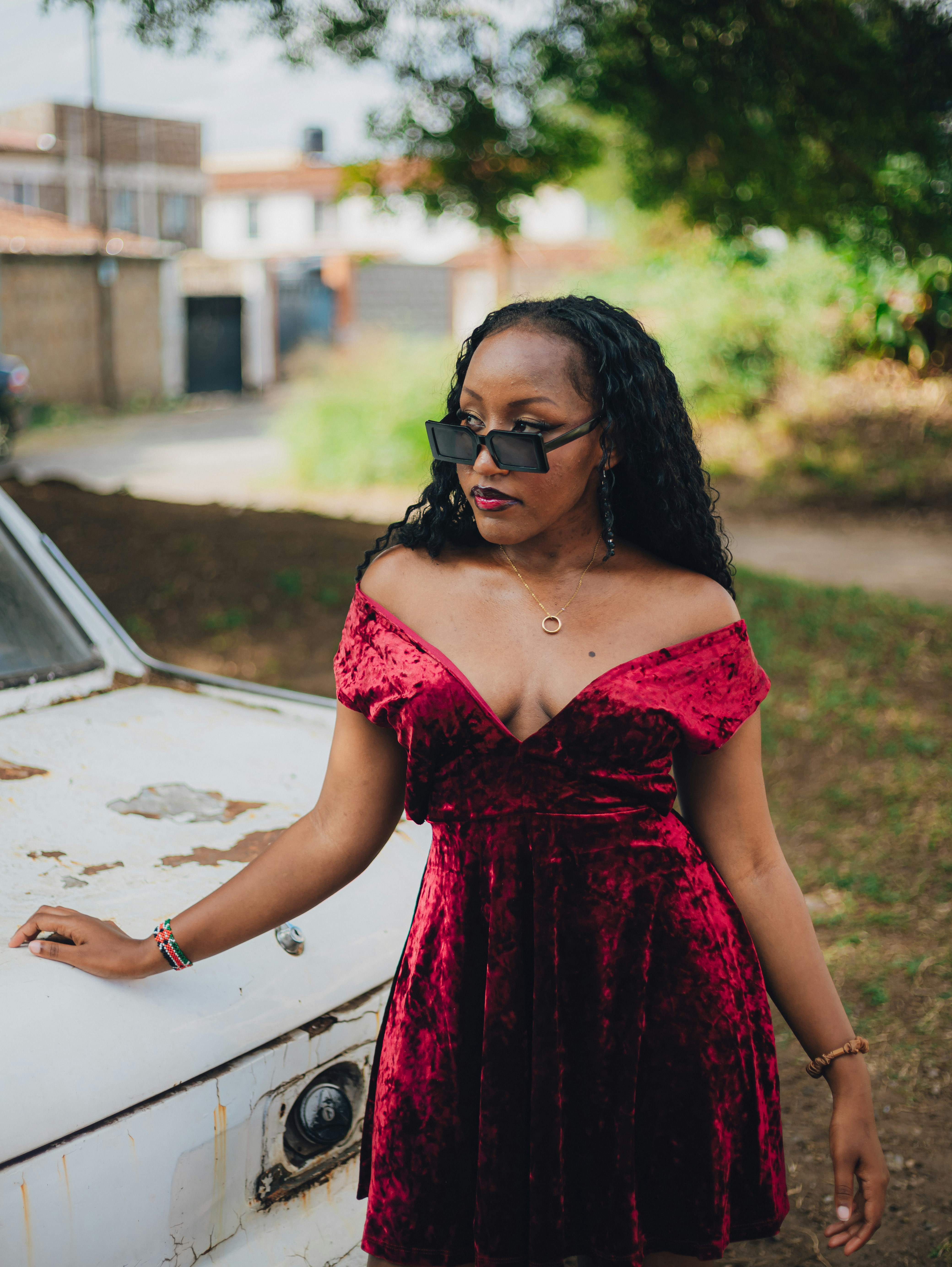 Young woman in red velvet dress poses by old car.
