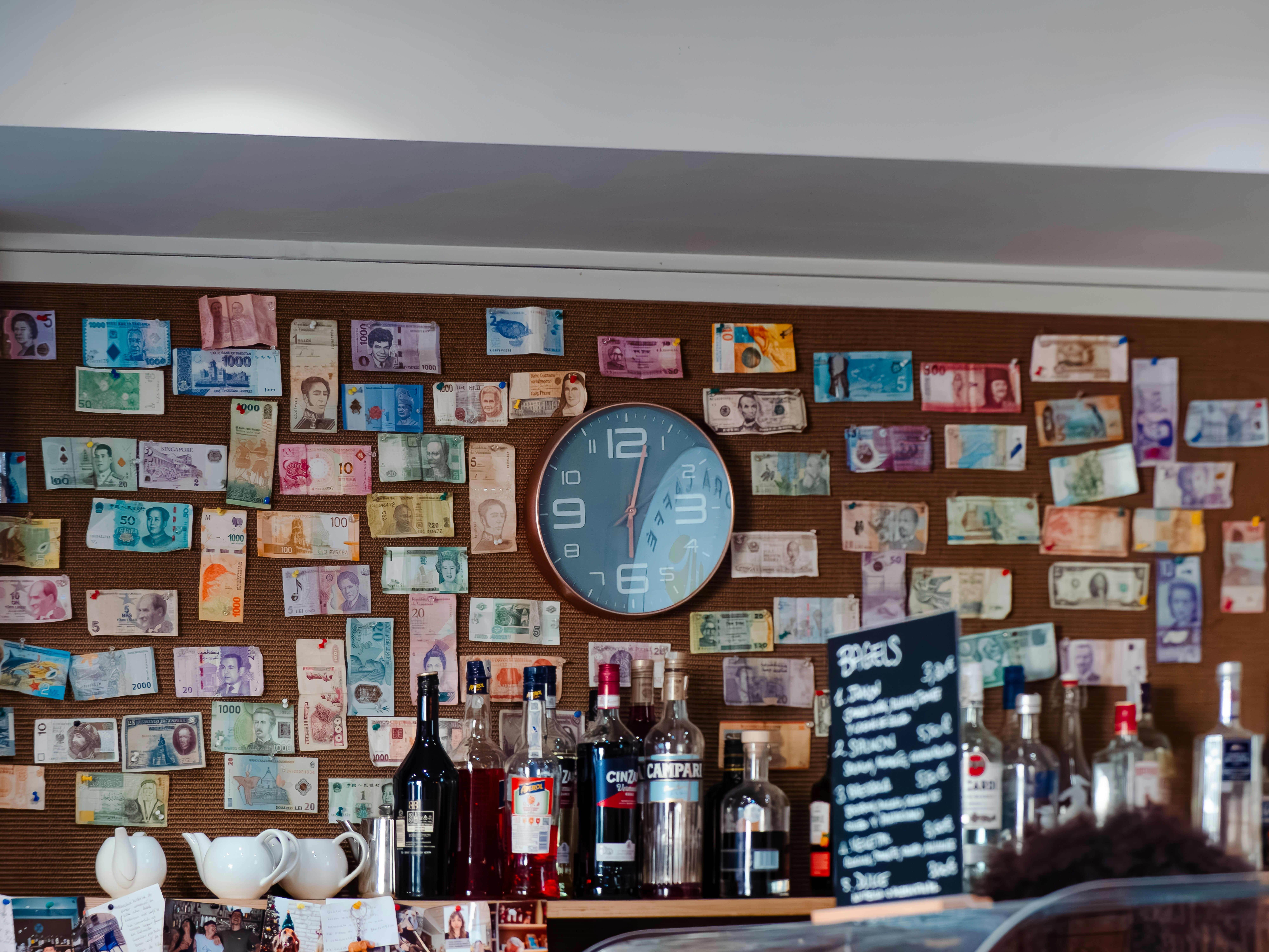 A wall covered in international banknotes with a round clock and liquor bottles