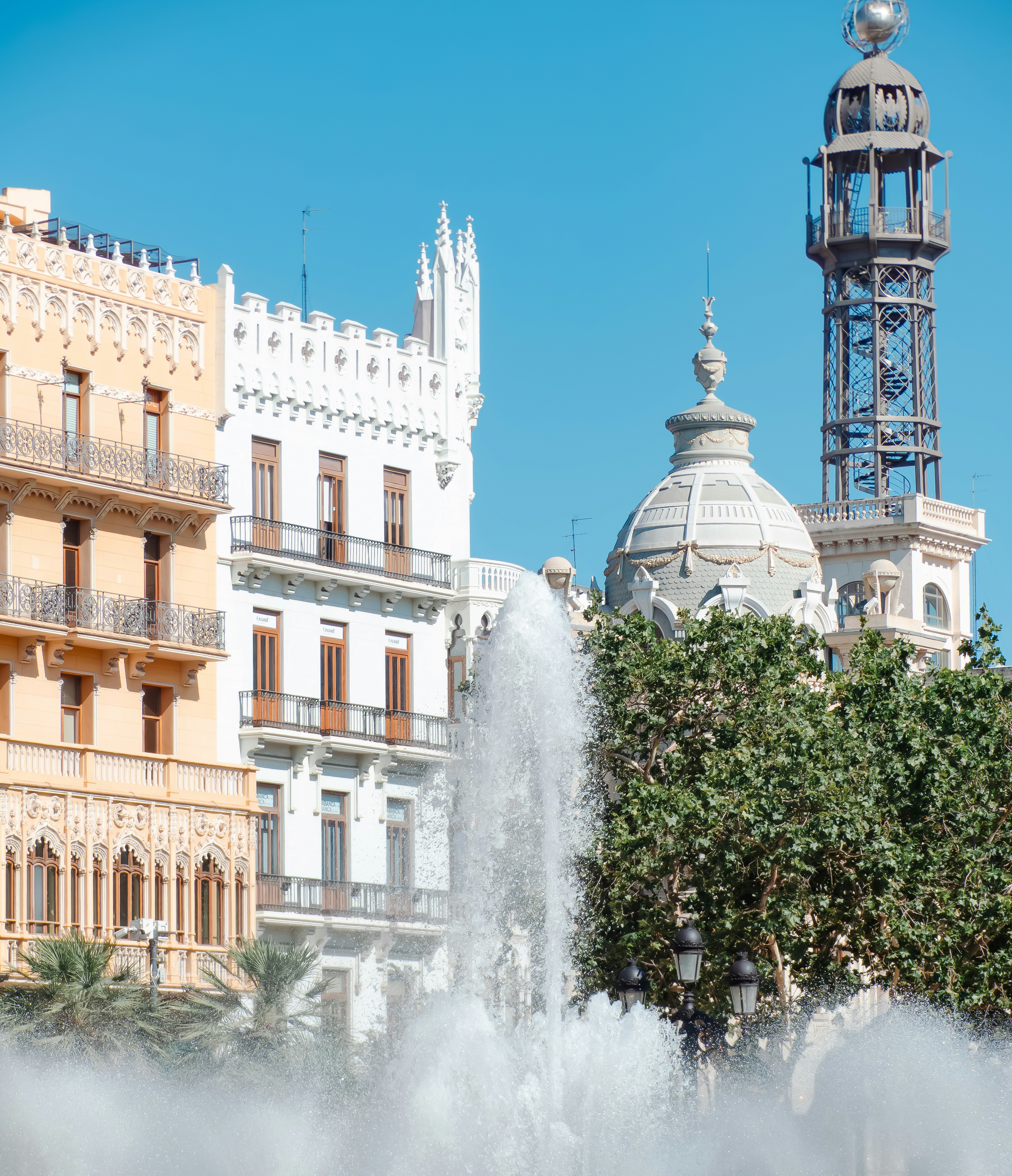 A fountain spraying water before ornate buildings and a tall metal tower in Valencia