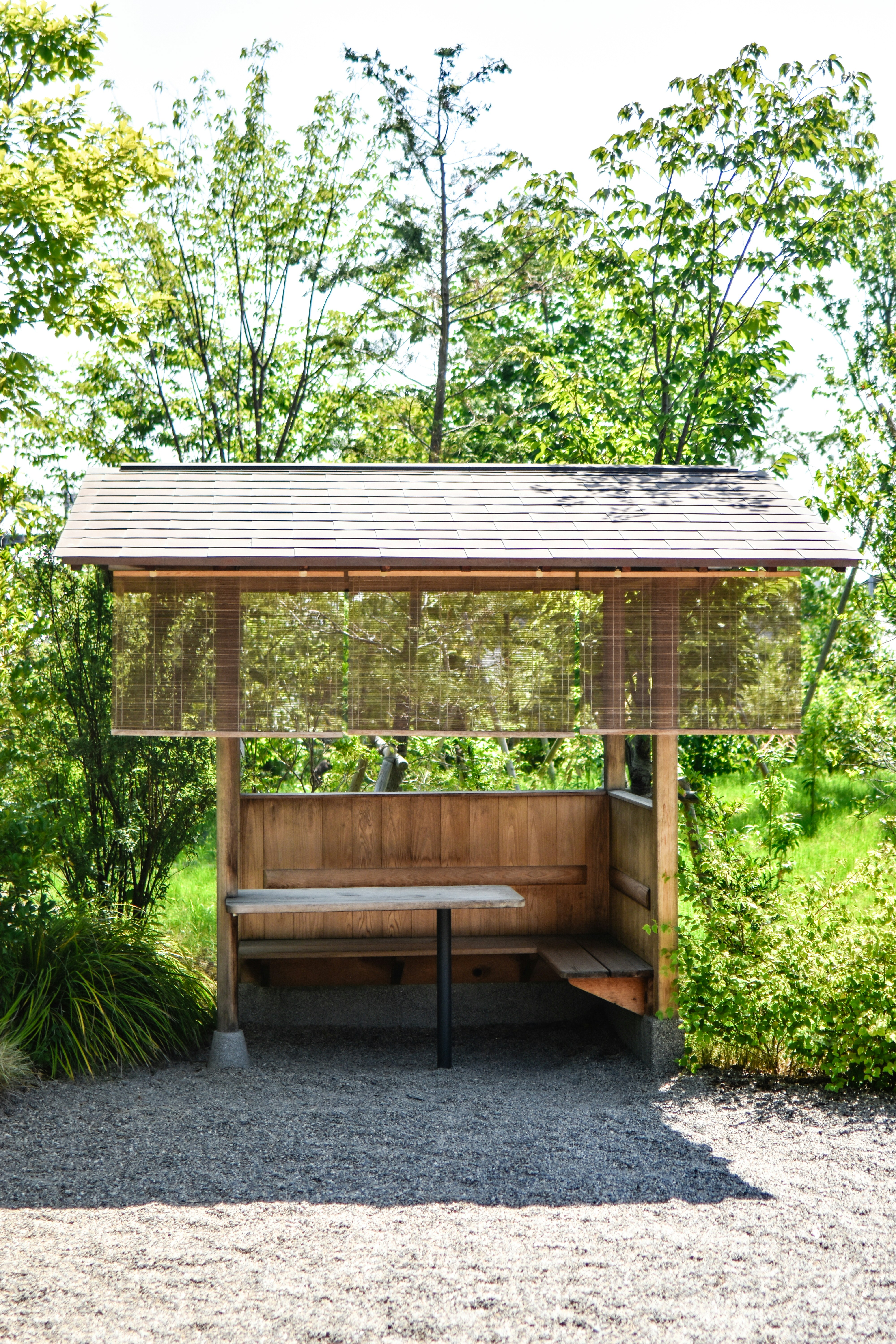 Tempat berlindung kayu luar ruangan dengan bangku dan meja yang dikelilingi oleh pepohonan hijau.