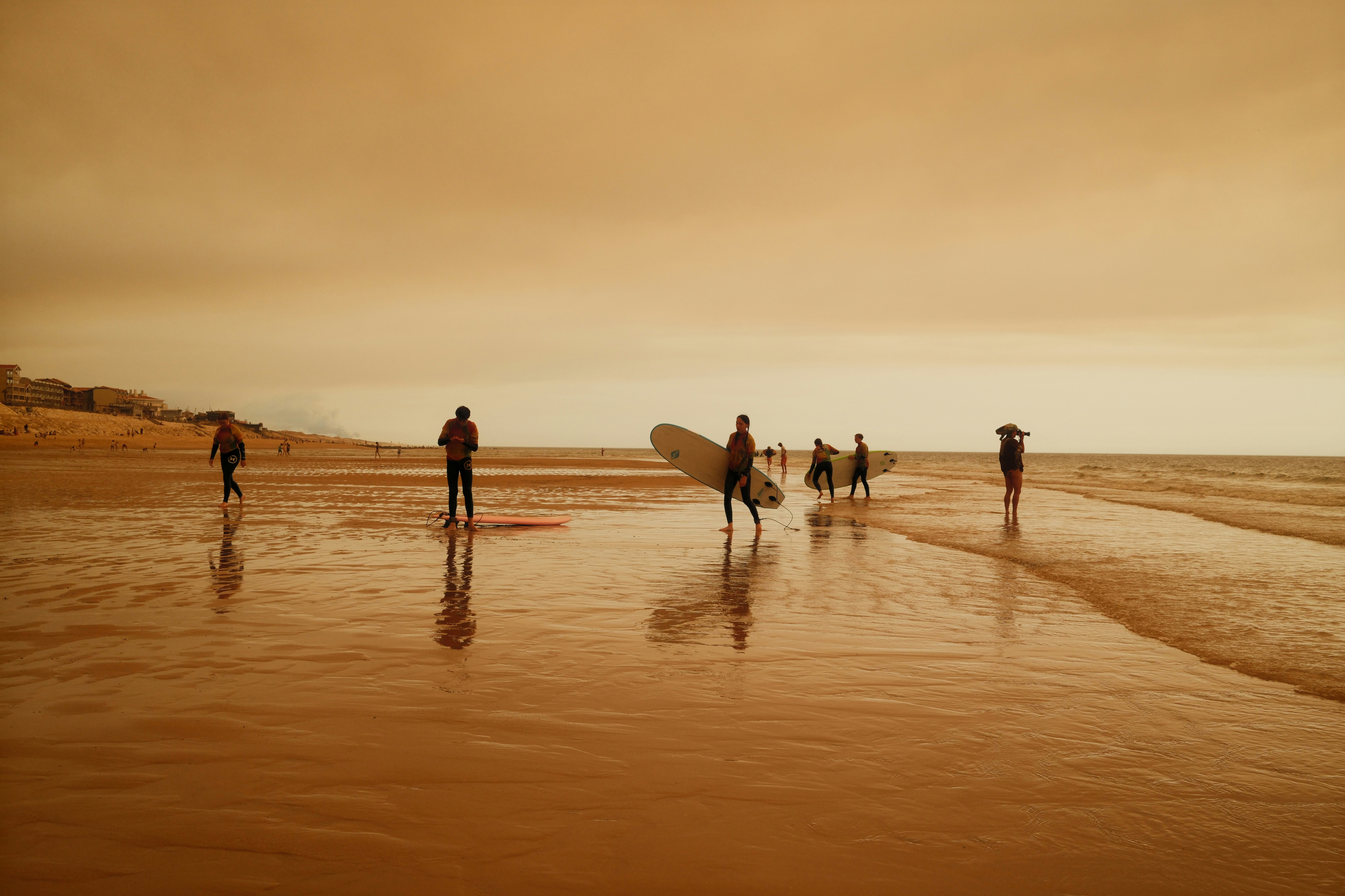 Des surfeurs avec des planches sur une plage de sable mouillée sous un ciel orange brumeux
