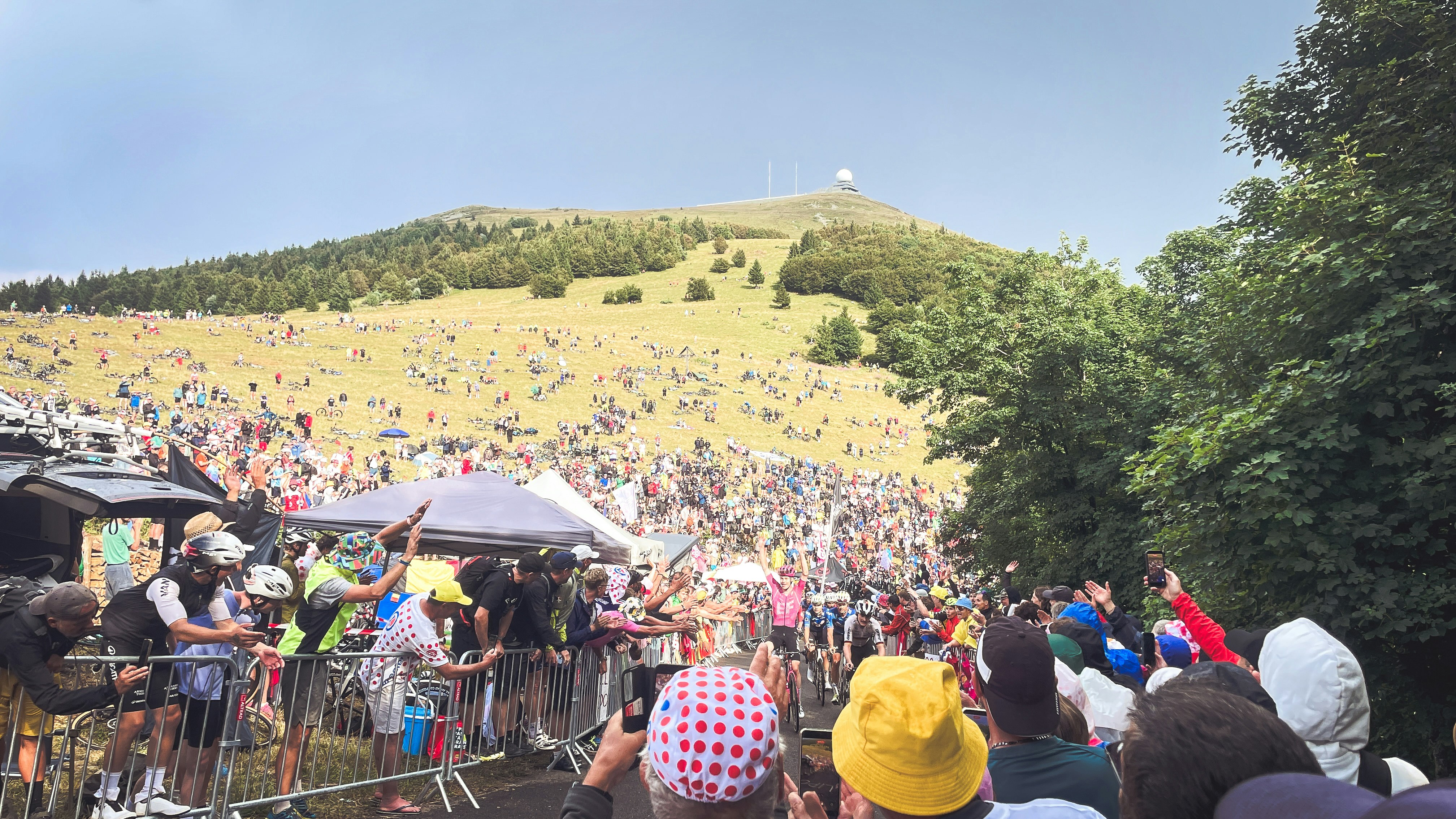 A large crowd watches an event near a grassy mountain.