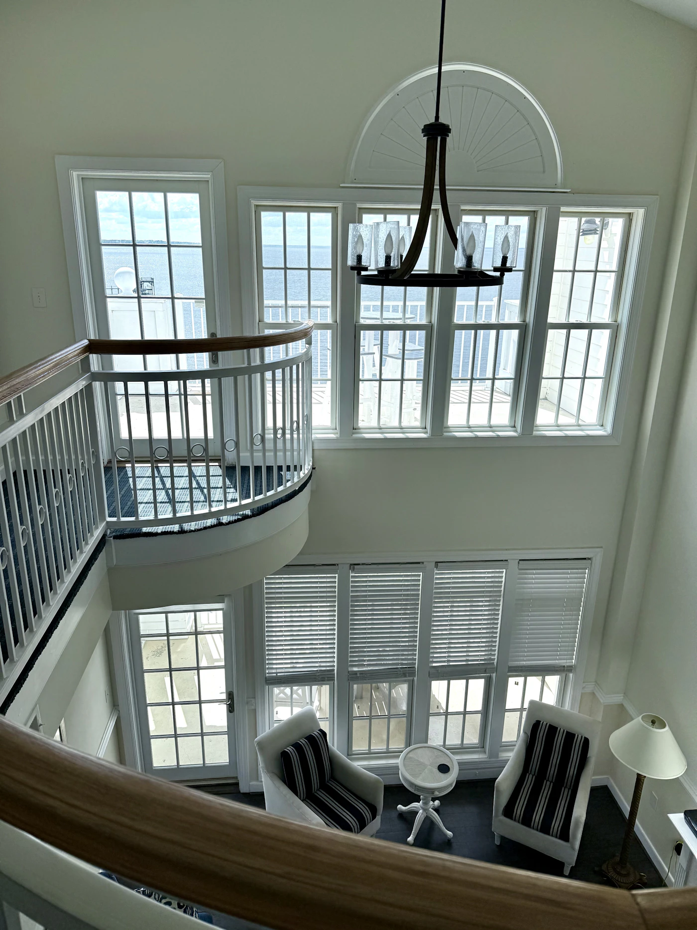 Bright double-height coastal interior with extensive windows overlooking the water, a curved staircase railing, a hanging chandelier and a small seating area below