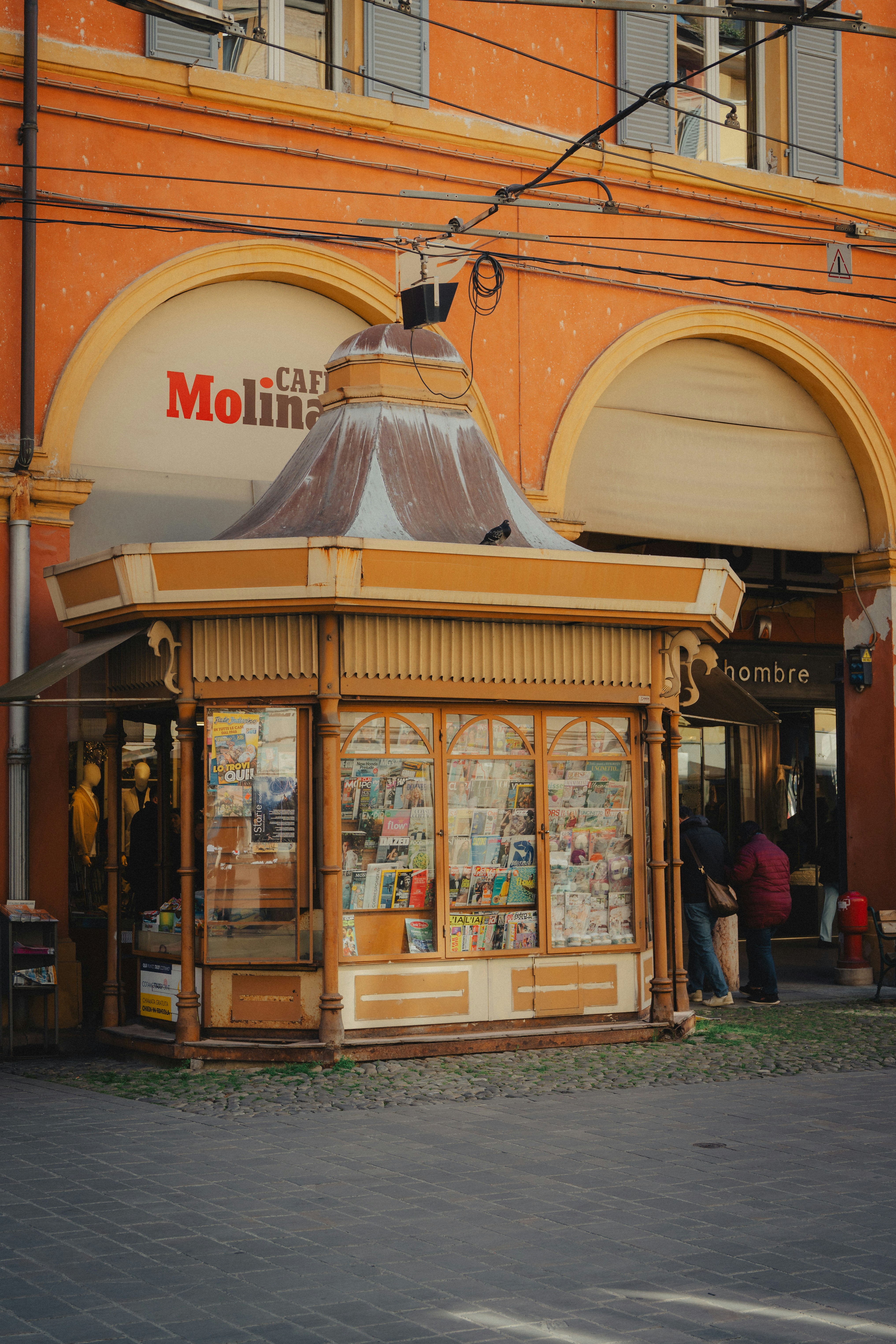 A vintage newsstand with glass windows displaying magazines in a sunlit city square.