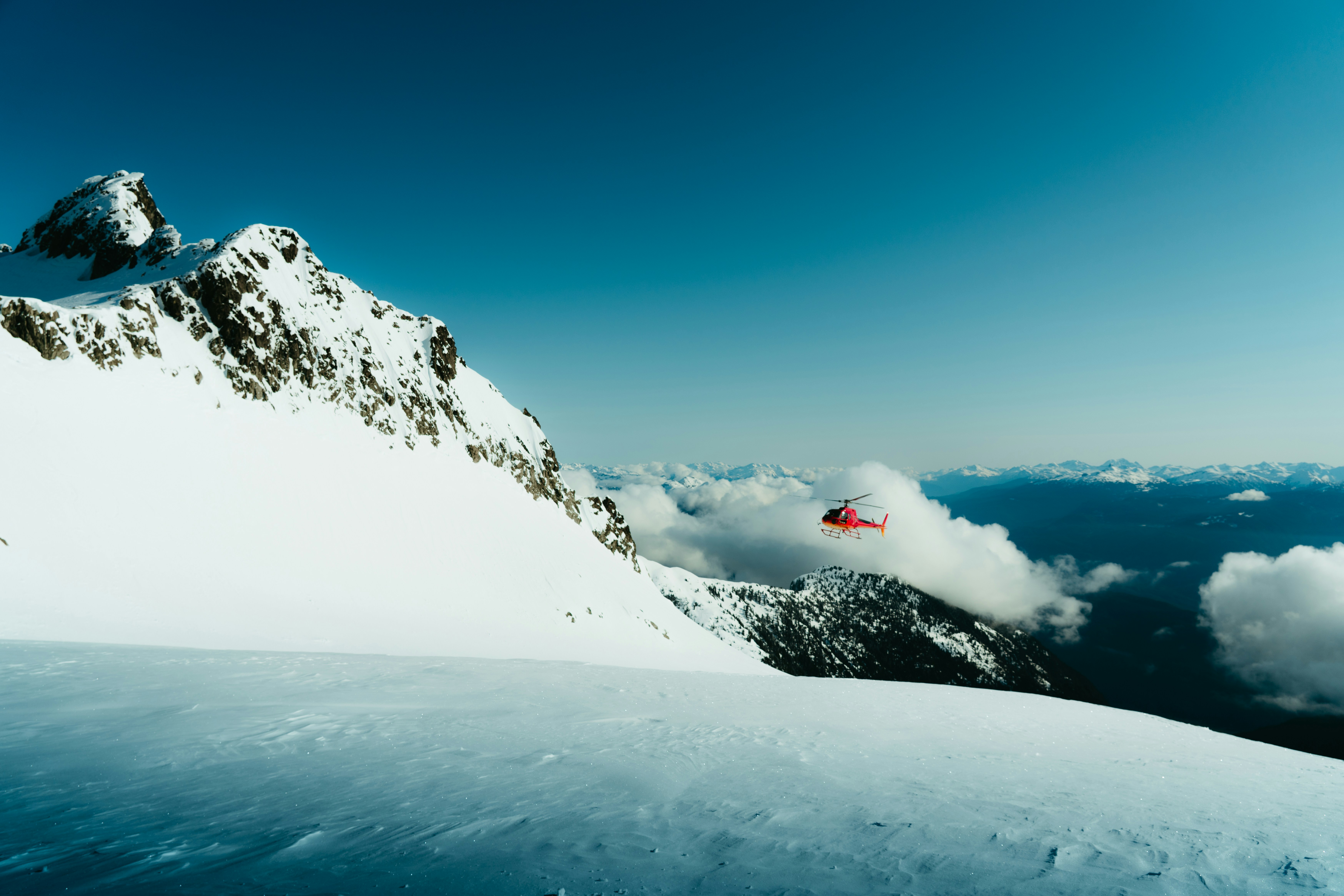 Red helicopter flying near snow-capped mountains under a blue sky.